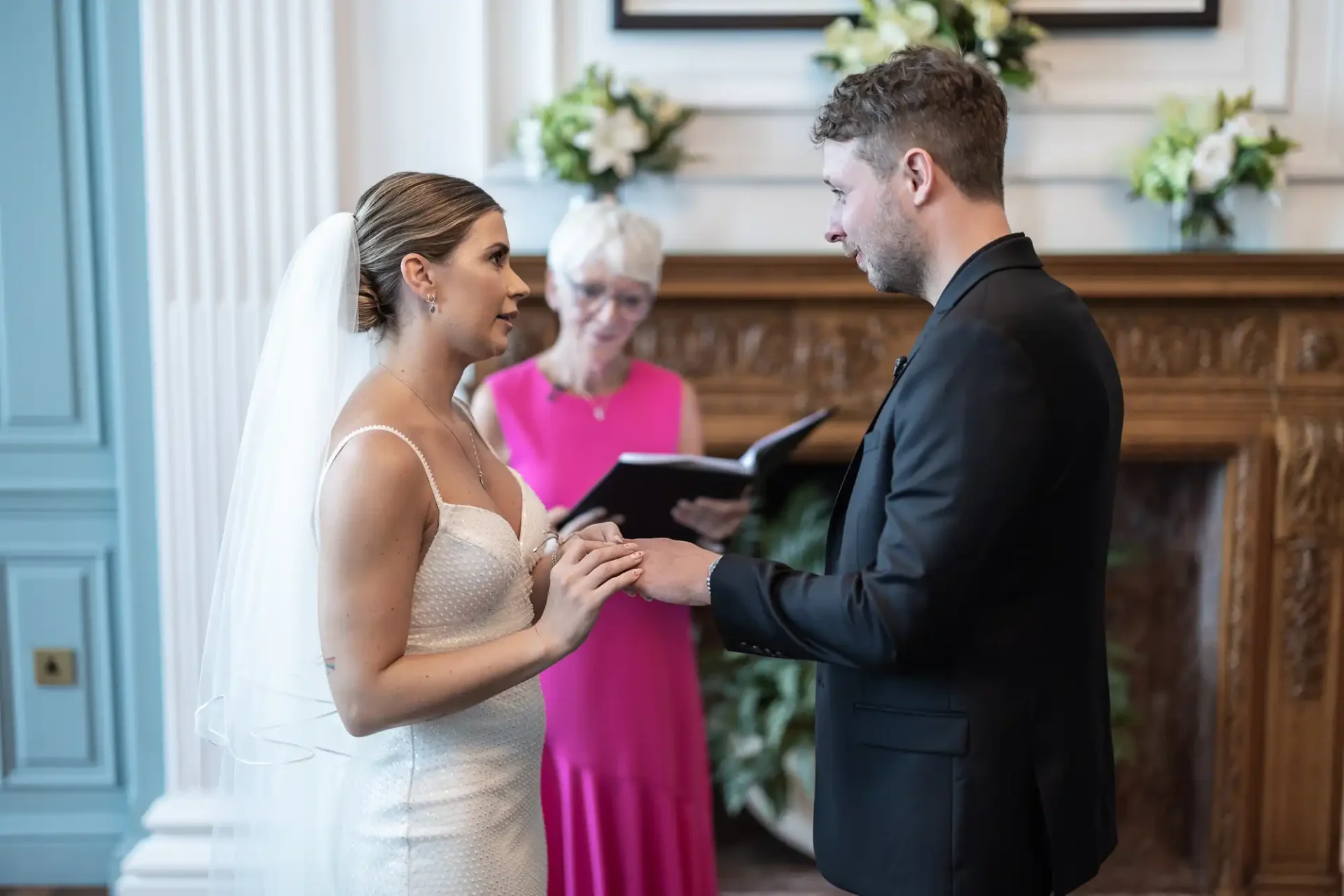 A bride and groom exchange vows in a sophisticated room with an officiant in pink reading from a book.