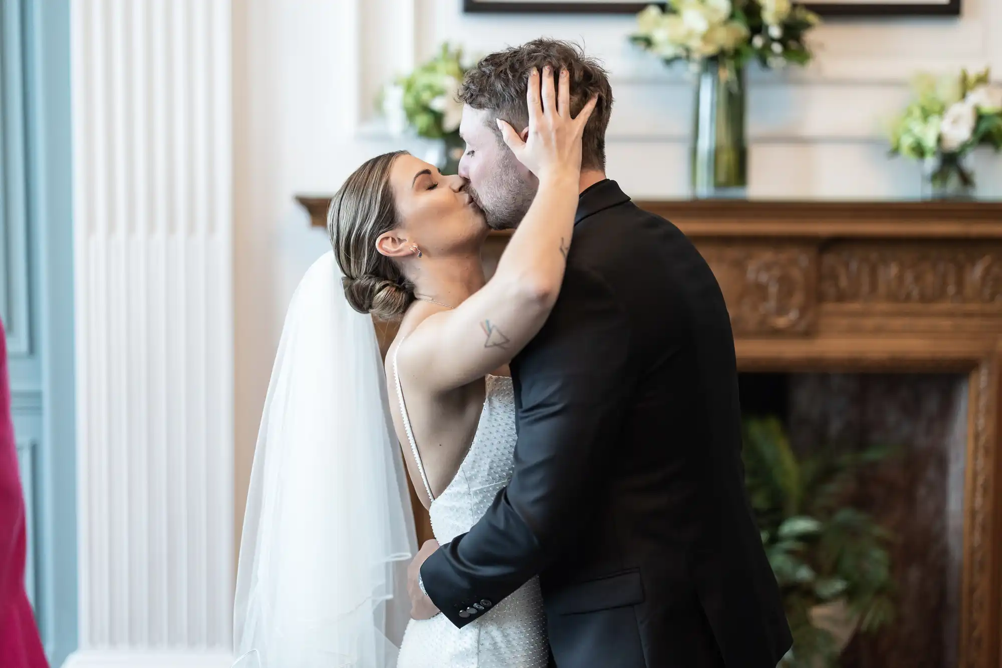 A bride and groom share a kiss in front of a fireplace adorned with flowers at their wedding ceremony.