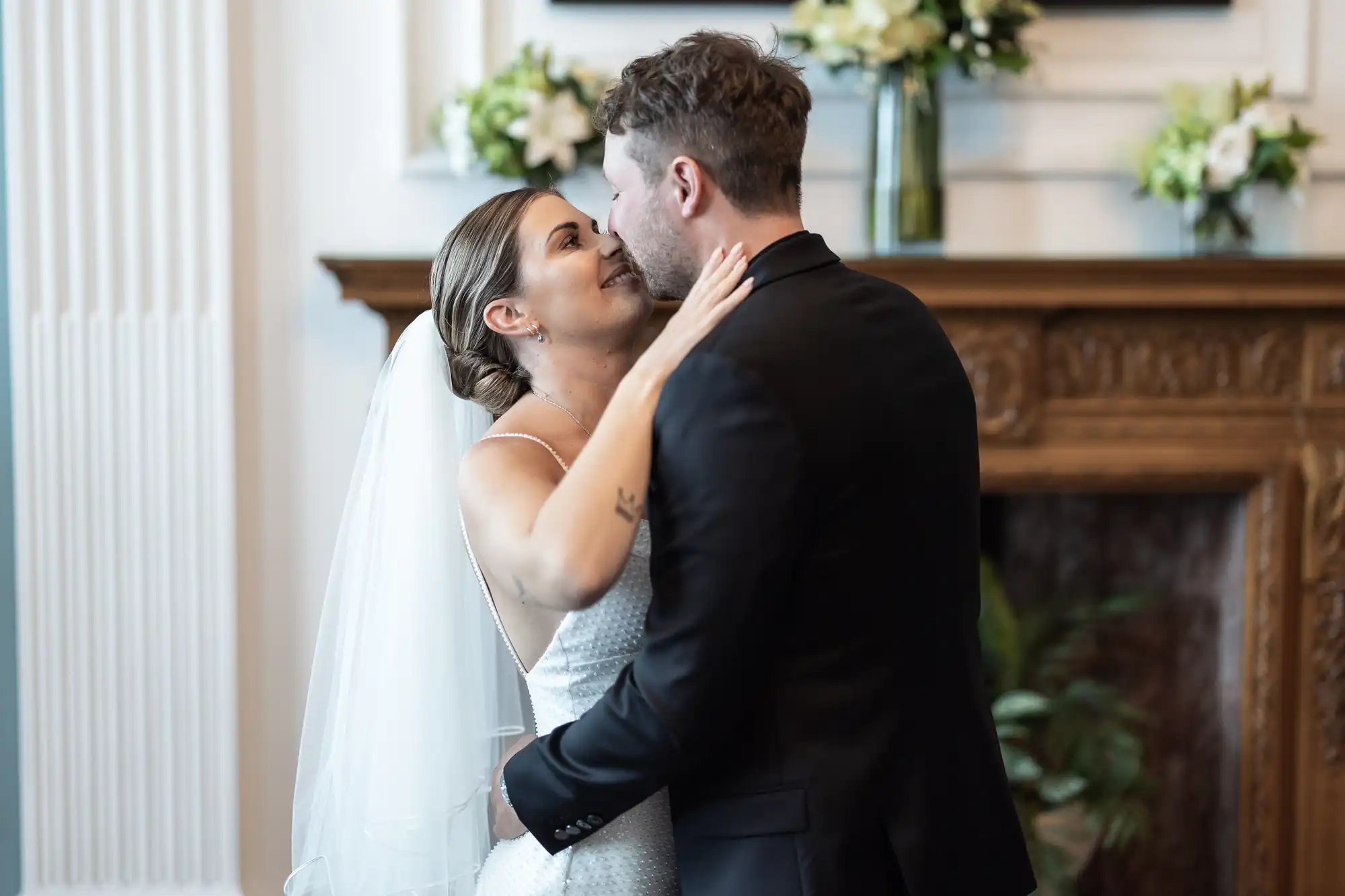 A bride and groom share a kiss in front of a fireplace decorated with flowers at their wedding ceremony.