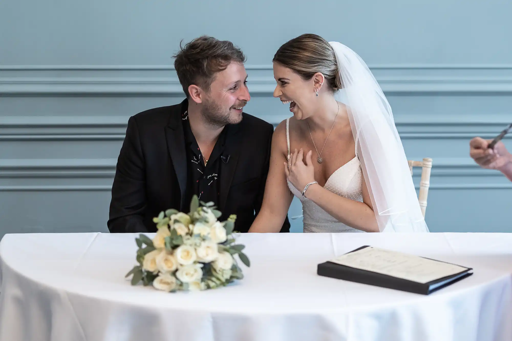 A newlywed couple happily interacting at a signing table with a bouquet and a guestbook, in a room with light blue walls.