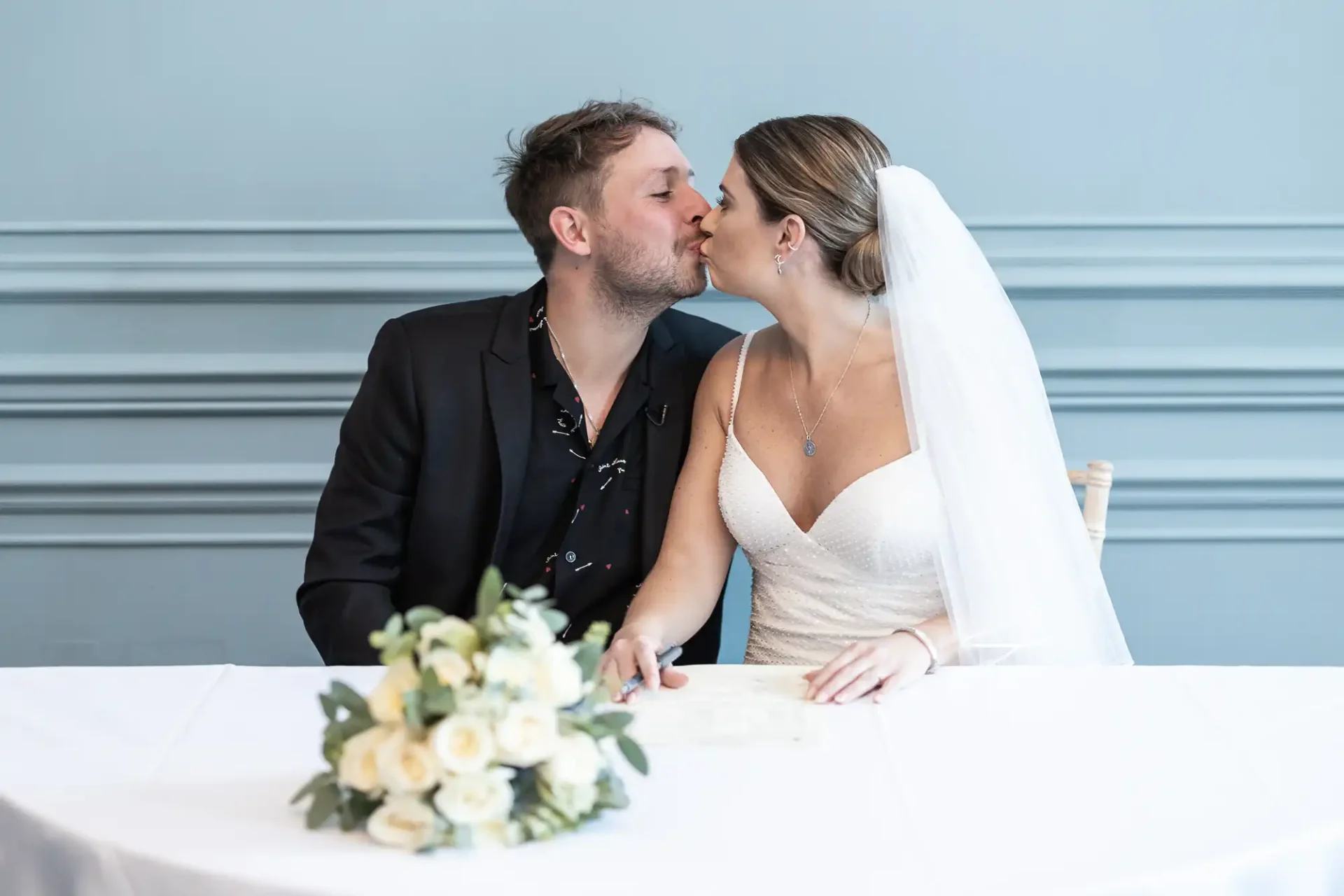 A bride and groom kissing at a signing table with a bouquet of white roses in the foreground.