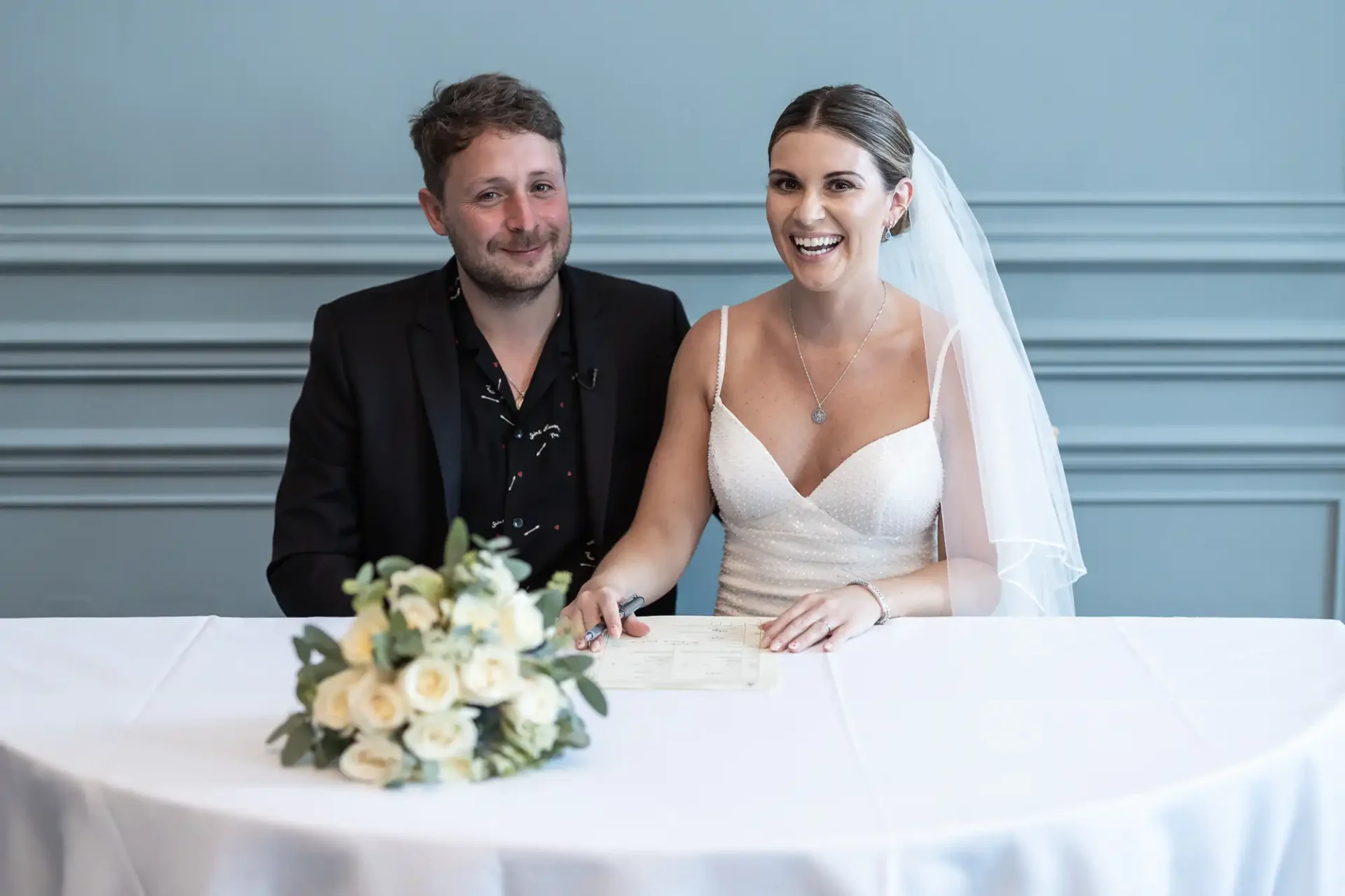 A happy couple in wedding attire signing their marriage certificate at a table, with a bouquet of flowers in the foreground.