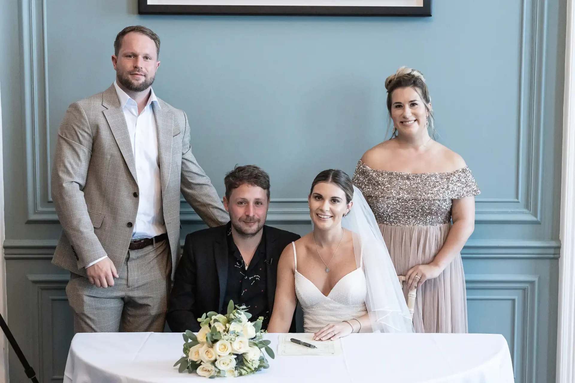 A bride and groom seated at a signing table, flanked by a best man and a bridesmaid, in a room with light blue walls.