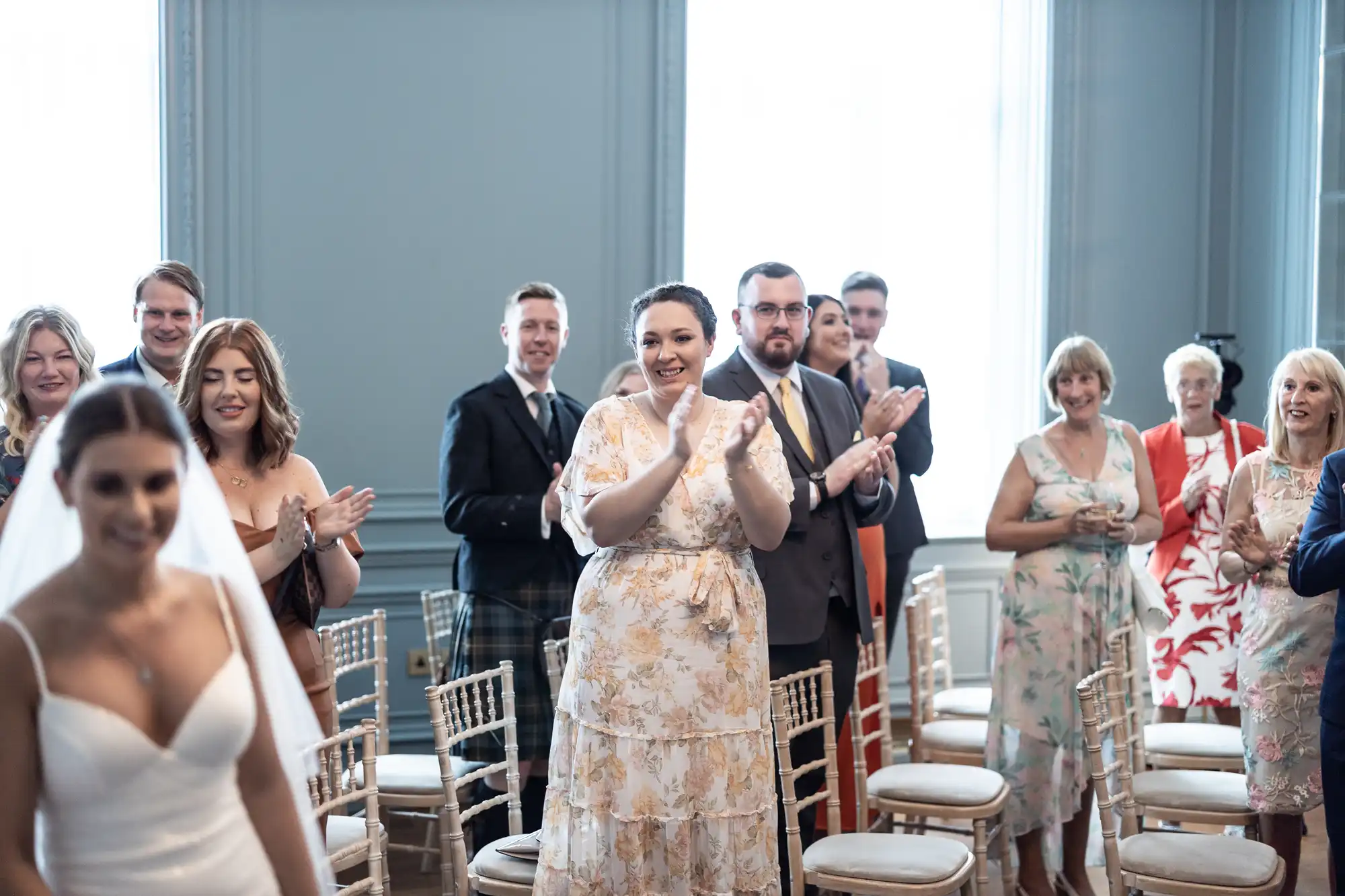 A bride walking down the aisle as guests, including a woman in a floral dress clapping enthusiastically, look on with smiles in a brightly lit room.