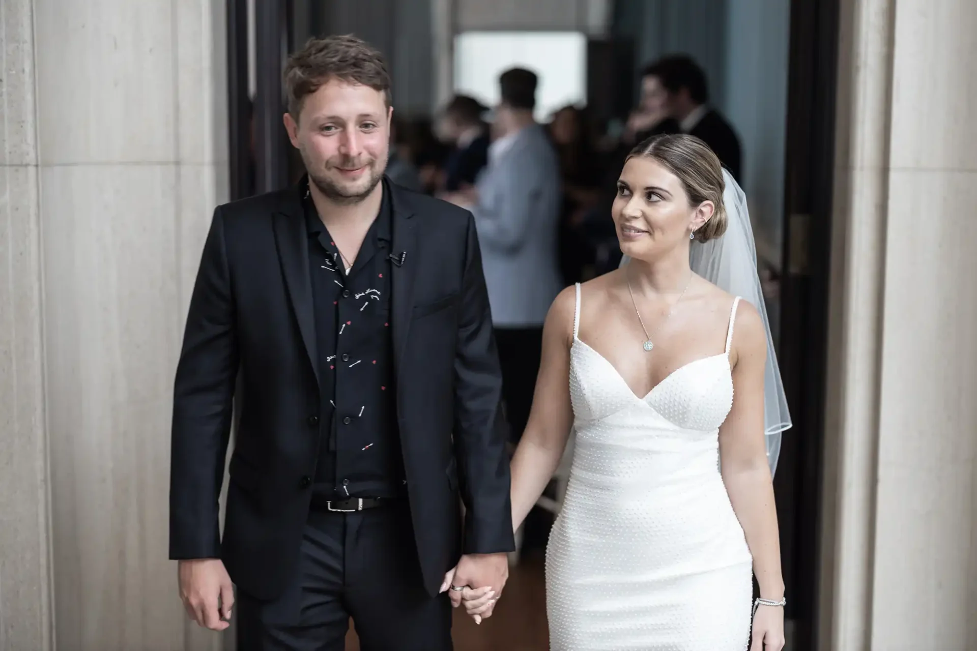 A bride in a white gown and a groom in a black suit walk hand in hand with smiles, indoors at a wedding venue.