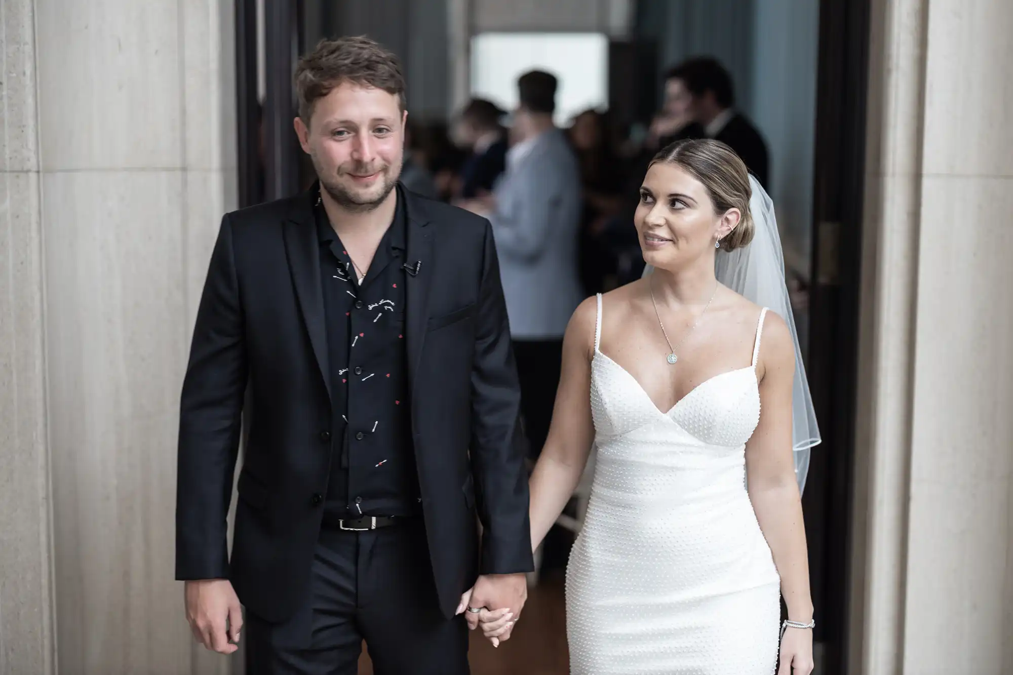 A bride in a white gown and a groom in a black suit walk hand in hand with smiles, indoors at a wedding venue.