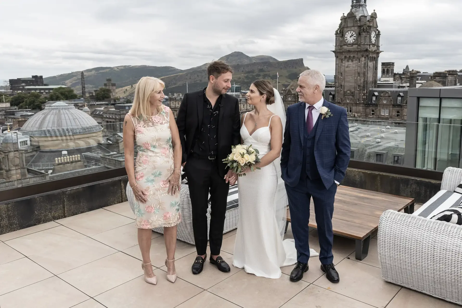 A newlywed couple with their parents on a rooftop, cityscape in the background, all smiling and dressed formally.