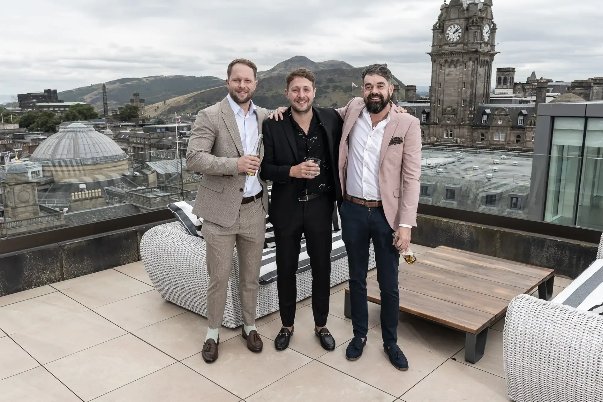 Three men in smart casual attire holding drinks on a rooftop terrace with cityscape and cloudy sky in the background.