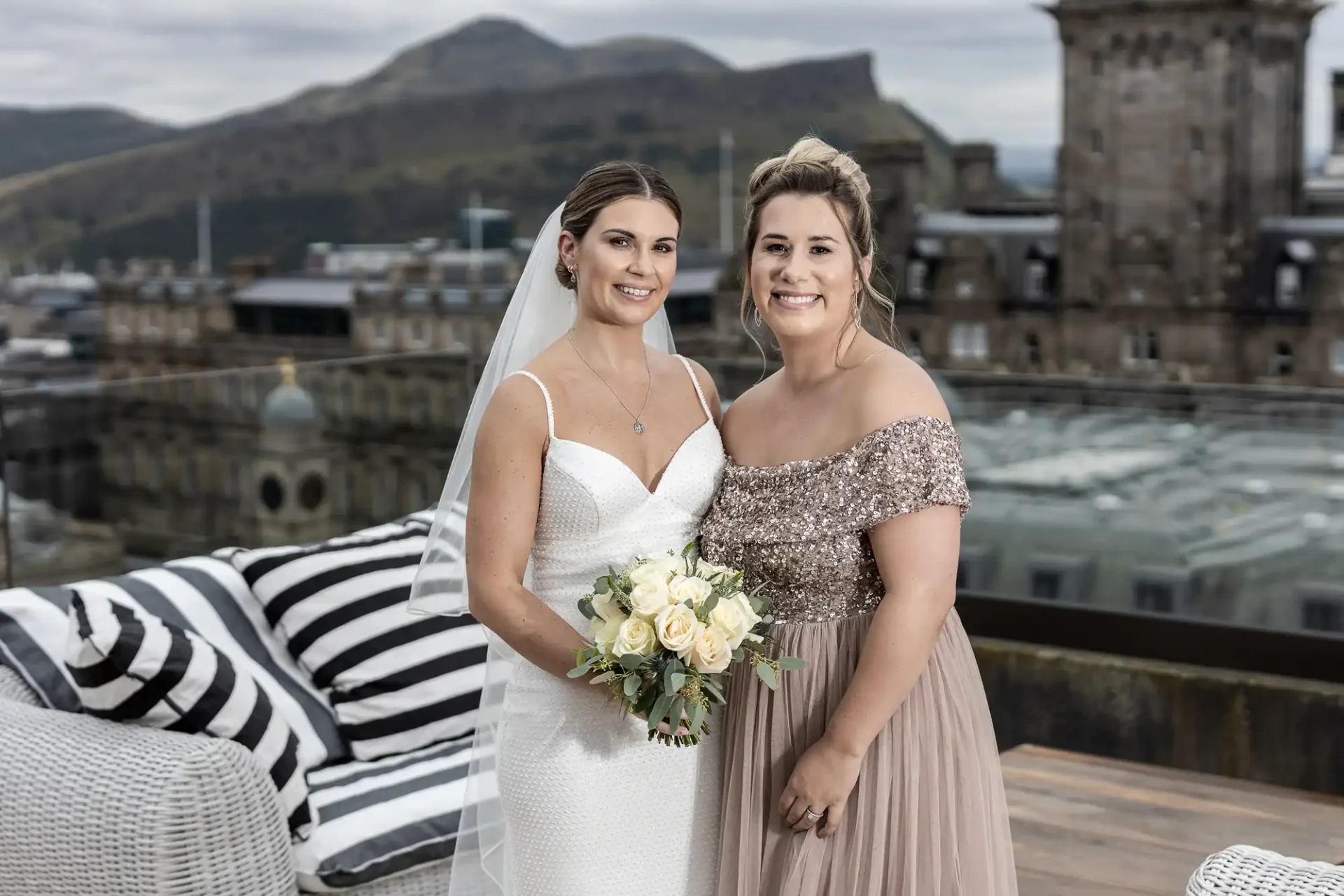 Two women smiling on a rooftop, one in a white wedding dress holding a bouquet and the other in a sequined beige dress, with city hills in the background.