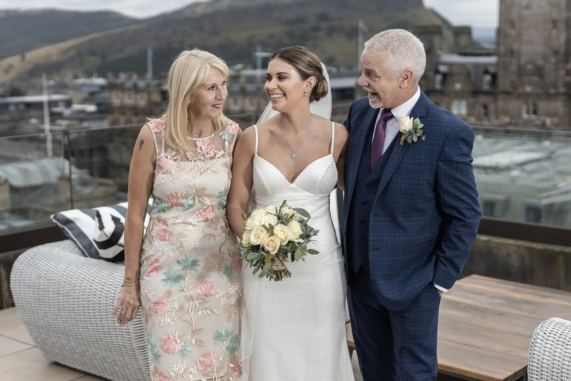 A bride stands smiling with her parents on a rooftop, cityscape in the background, all dressed in formal wear.