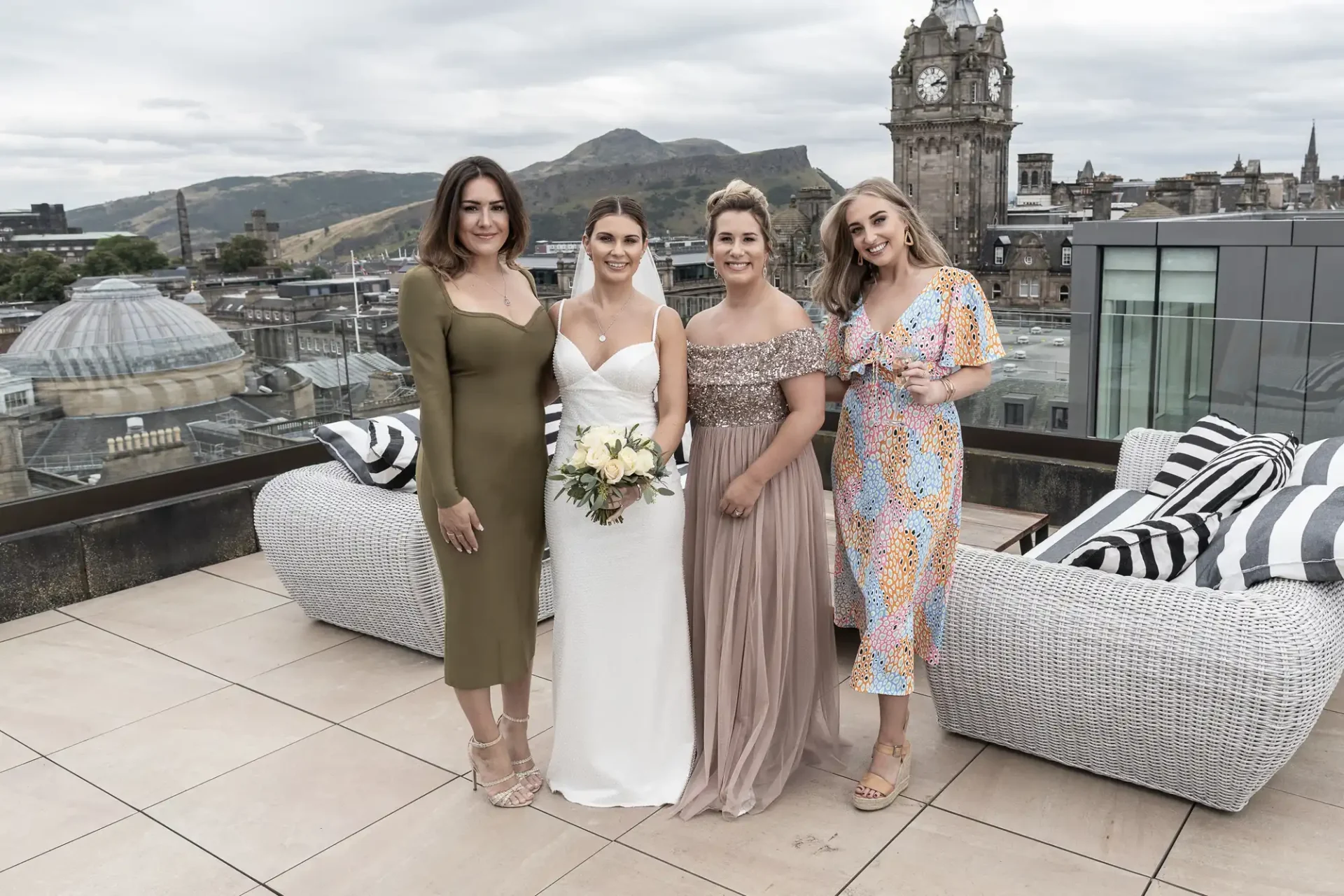 Four women in formal dresses posing together on a rooftop with a cityscape background.