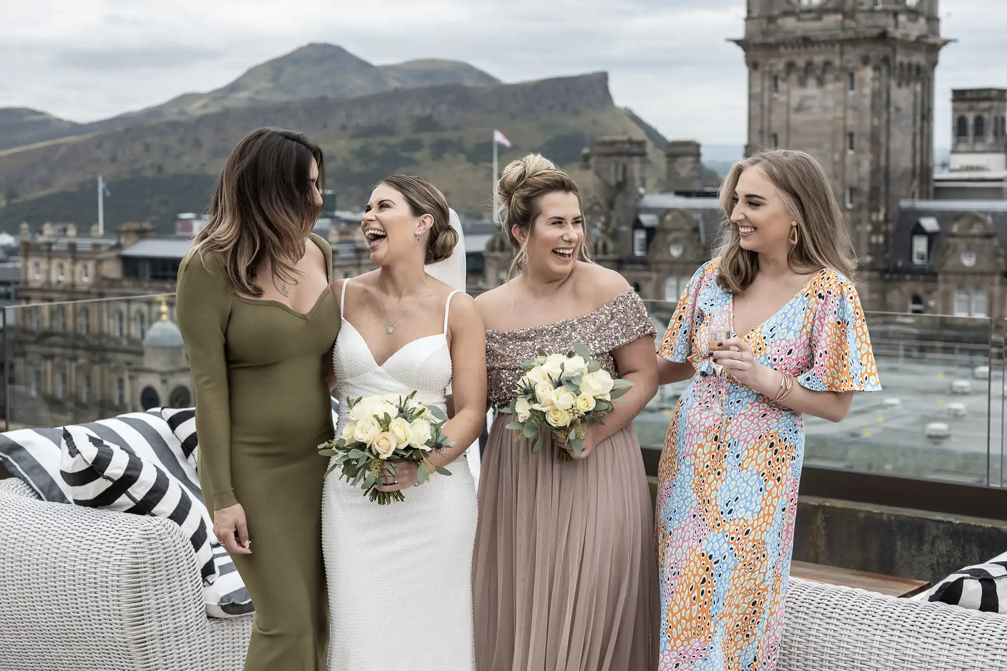 Four women in formal dresses, holding bouquets and laughing on a rooftop with a cityscape and hills in the background.