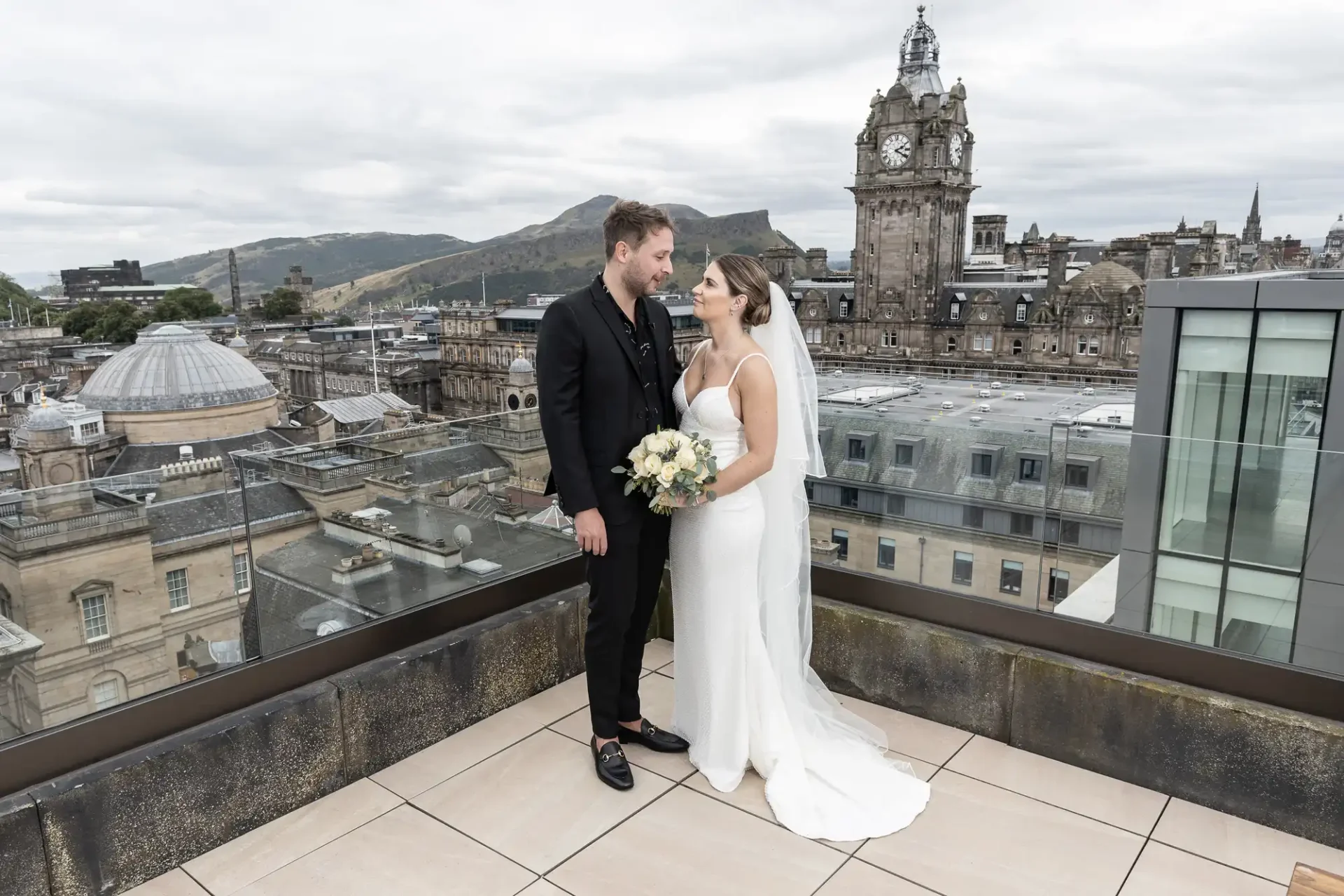 A bride and groom standing on a rooftop, affectionately looking at each other, with an urban skyline in the background.