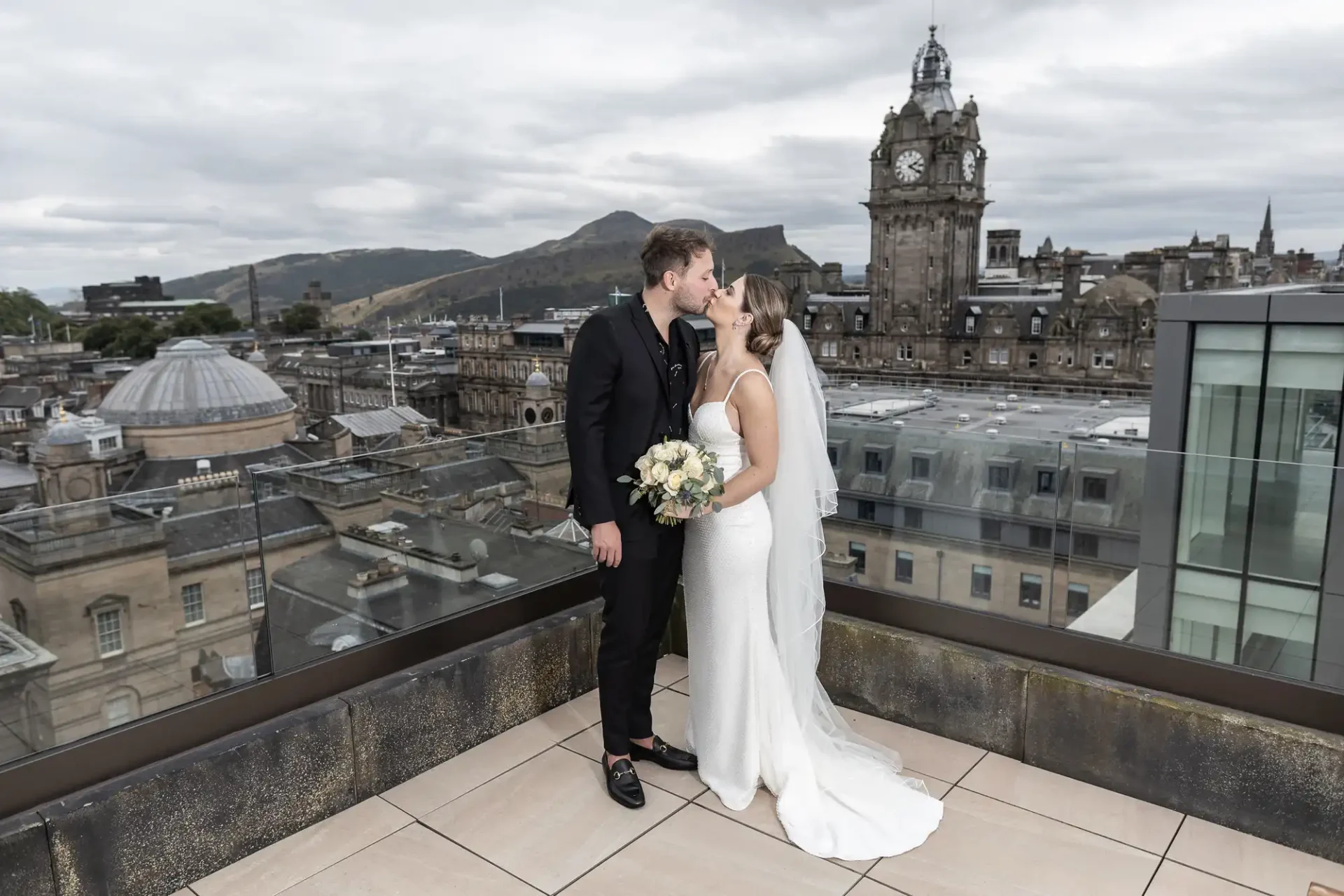 A bride and groom kissing on a rooftop, with edinburgh's cityscape and hills in the background.