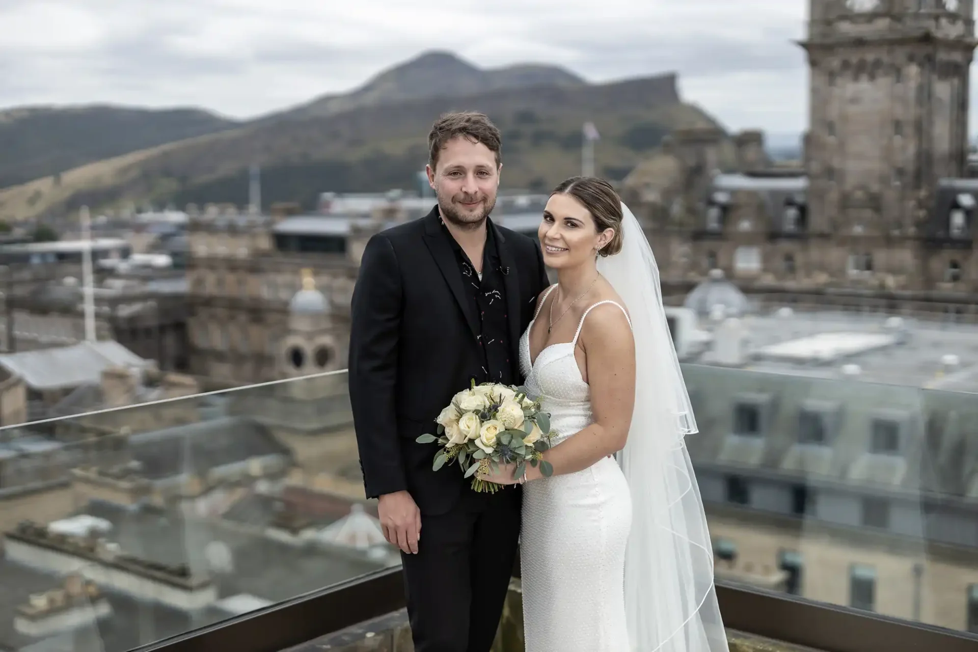 A bride and groom pose on a terrace with a cityscape and hills in the background, the bride holding a bouquet.