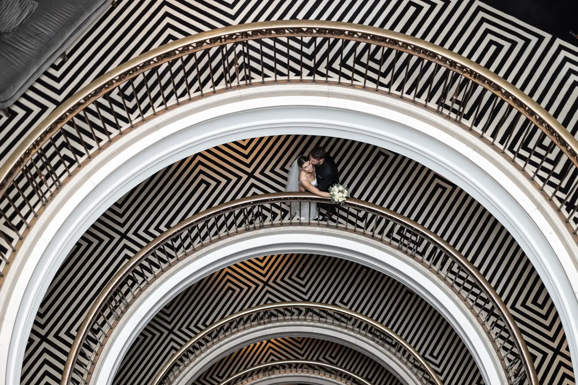 A bride and groom embrace on a balcony with geometric black and white patterns on the floors of a spiraling staircase.