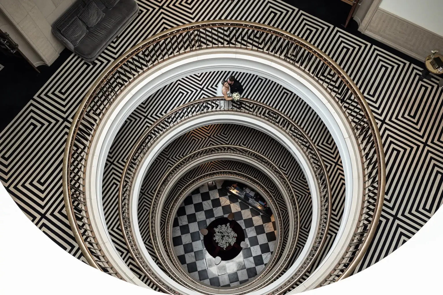 Overhead view of a person standing on a circular staircase with intricate black and white geometric patterns, leading down into a tiled foyer.