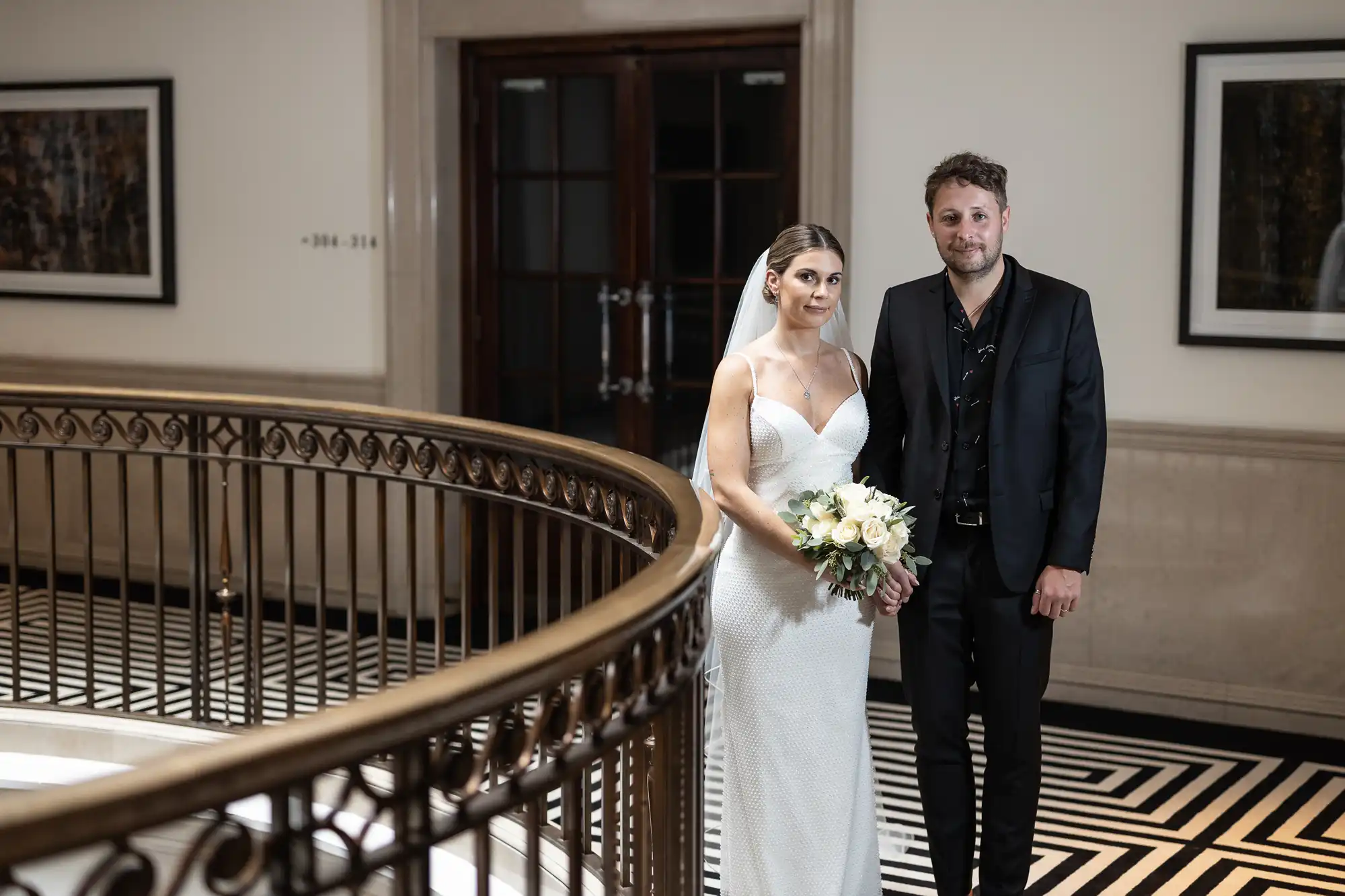 A bride in a white gown and a groom in a black suit stand together on a curved balcony, holding hands, with a backdrop of elegant doorways and framed paintings.