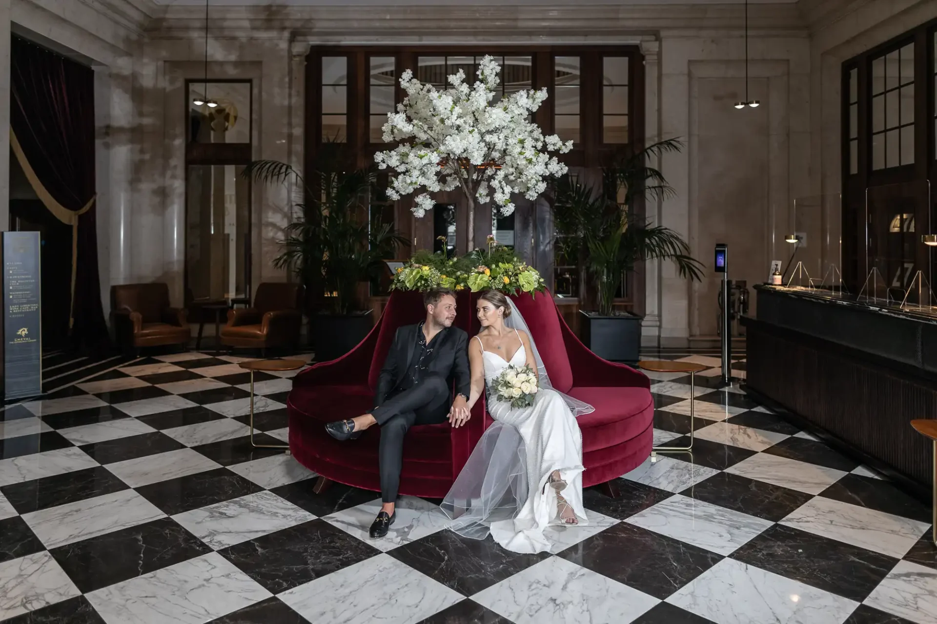 A bride and groom sitting together on a red sofa in a grand lobby with black and white tiled floors and a large floral arrangement.