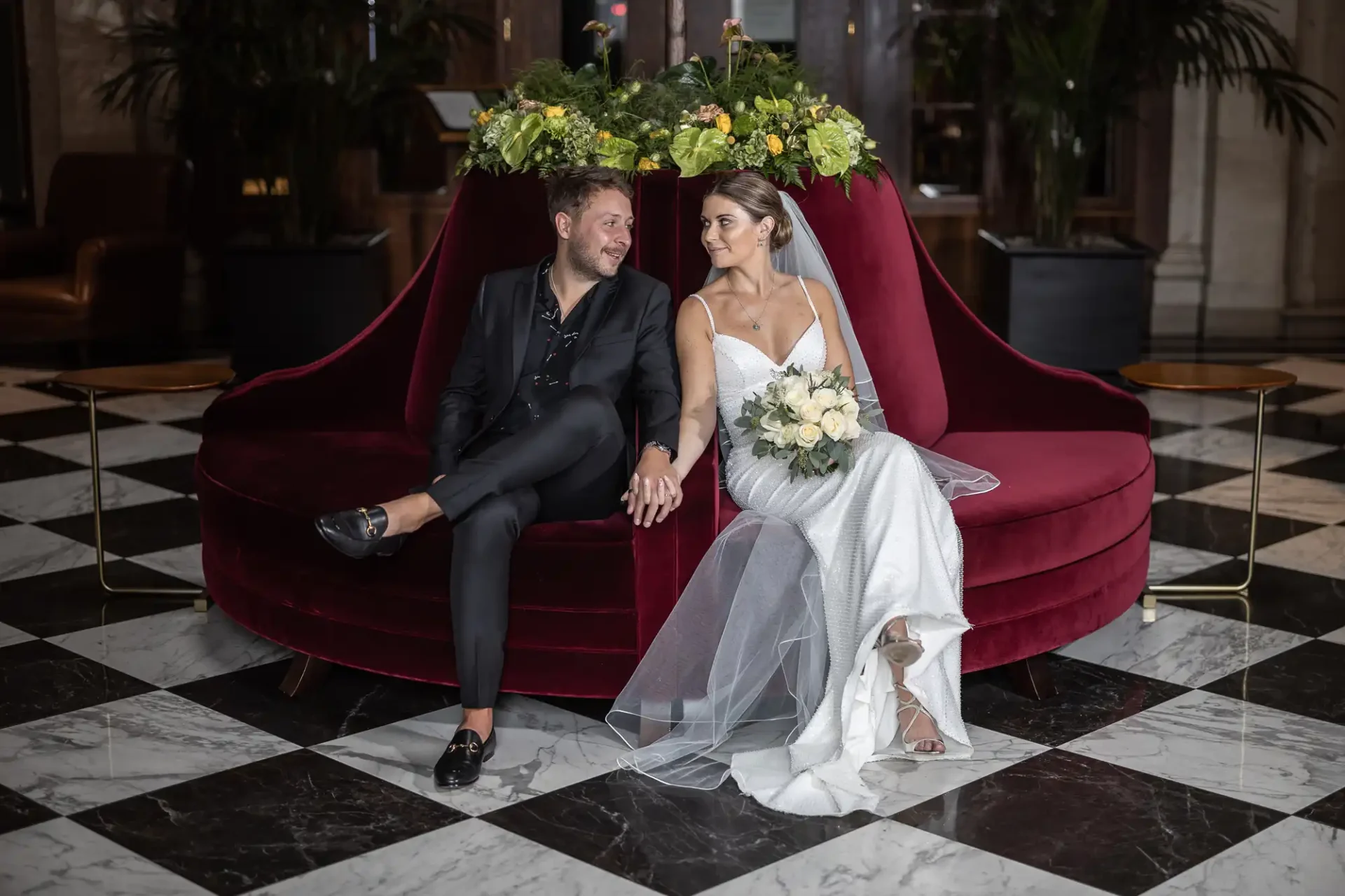 A newlywed couple, dressed in elegant wedding attire, sitting closely on a red velvet sofa, surrounded by flowers, in a luxurious lobby with black and white tiled flooring.