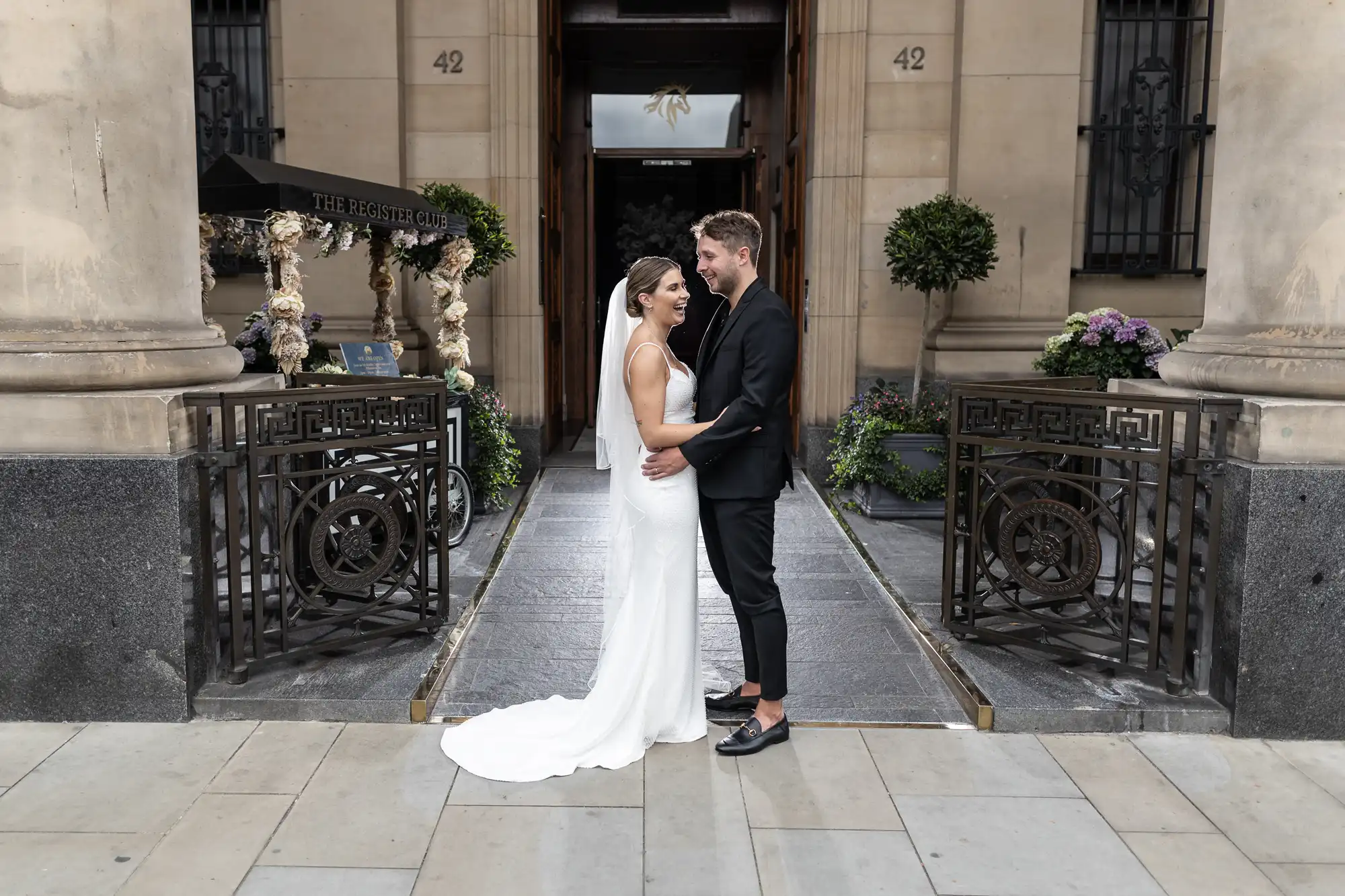 A newlywed couple stands lovingly in front of a building, with the bride in a white dress and the groom in a black suit.