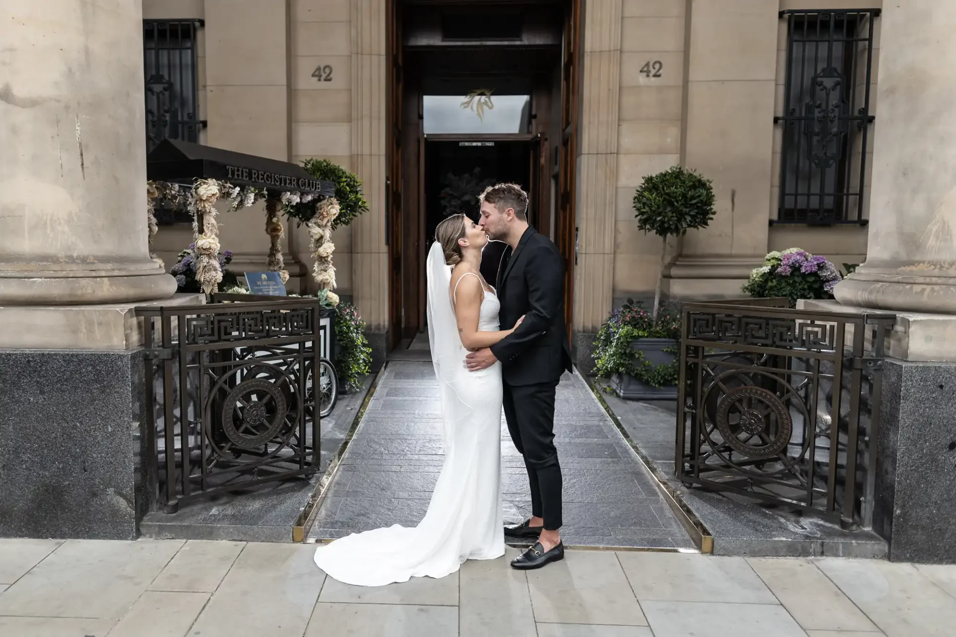 A bride in a white dress and a groom in a black suit kiss in front of a building entrance marked "42," adorned with floral decorations.