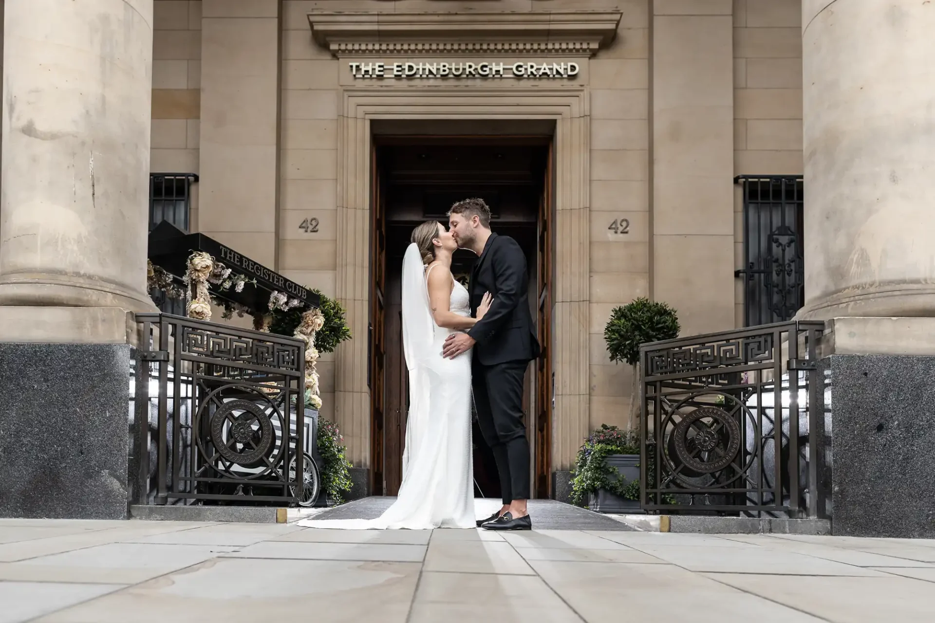 A couple kissing in front of "the edinburgh grand" entrance, framed by ornate black gates on a paved street.