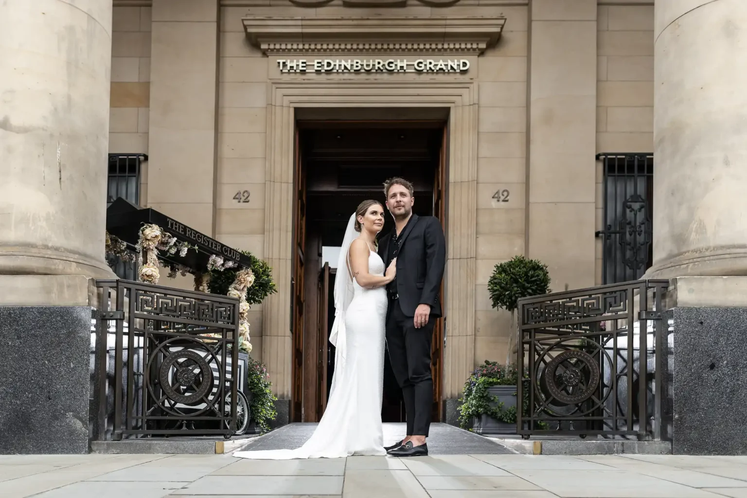 The Edinburgh Grand wedding photographer - luxury city centre wedding photography by Love Wedding Photos and Film. A bride in a white dress and veil stands beside a groom in a dark suit and sandals in front of The Edinburgh Grand entrance. They embrace, looking at the camera, surrounded by ornate railings, columns, and greenery on either side of the doorway.