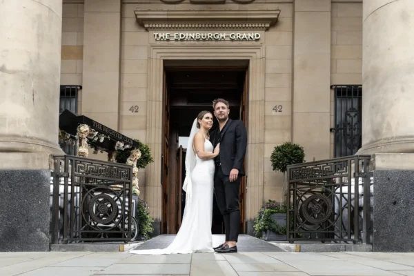 The Edinburgh Grand wedding photographer - luxury city centre wedding photography by Love Wedding Photos and Film. A bride in a white dress and veil stands beside a groom in a dark suit and sandals in front of The Edinburgh Grand entrance. They embrace, looking at the camera, surrounded by ornate railings, columns, and greenery on either side of the doorway.