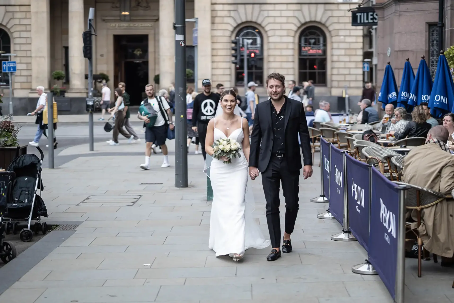 A newlywed couple walks hand in hand through a busy city street, the bride in a white dress with a bouquet, and the groom in a black suit.