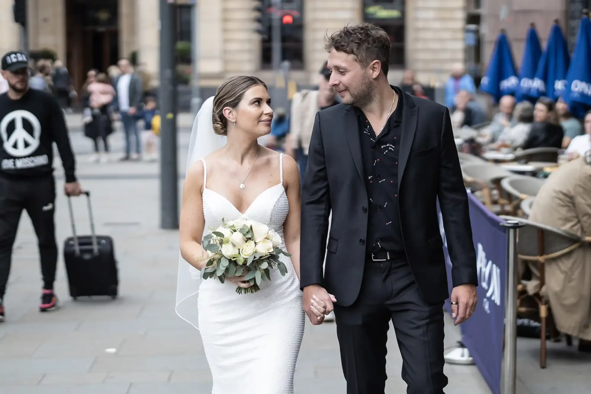 Newlywed couple walking hand in hand through a busy street, the bride holding a bouquet of white flowers, both smiling and looking at each other.