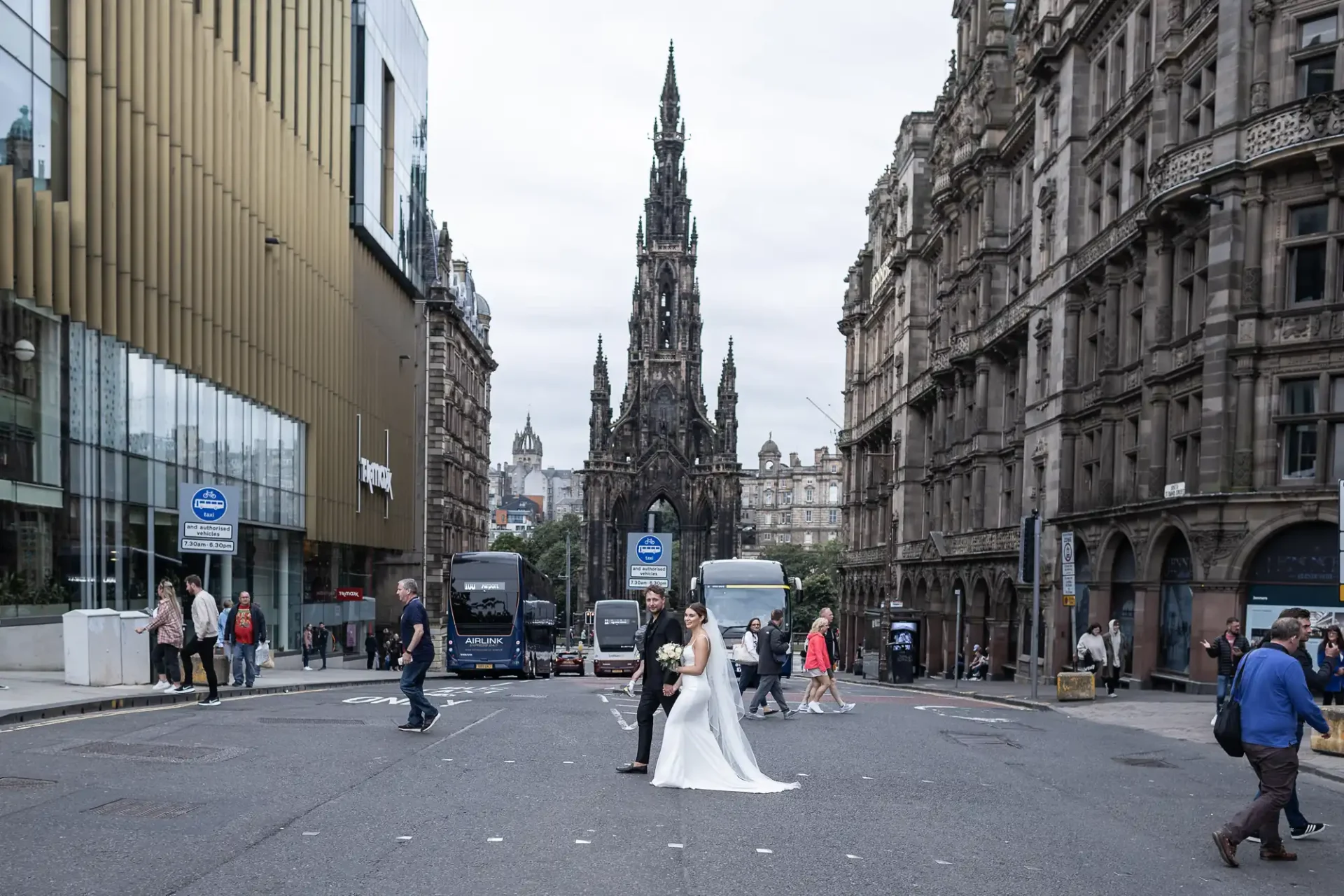 A bride and groom stand in the middle of a busy city street, posing for a photo with the scott monument in the background, surrounded by pedestrians and vehicles.