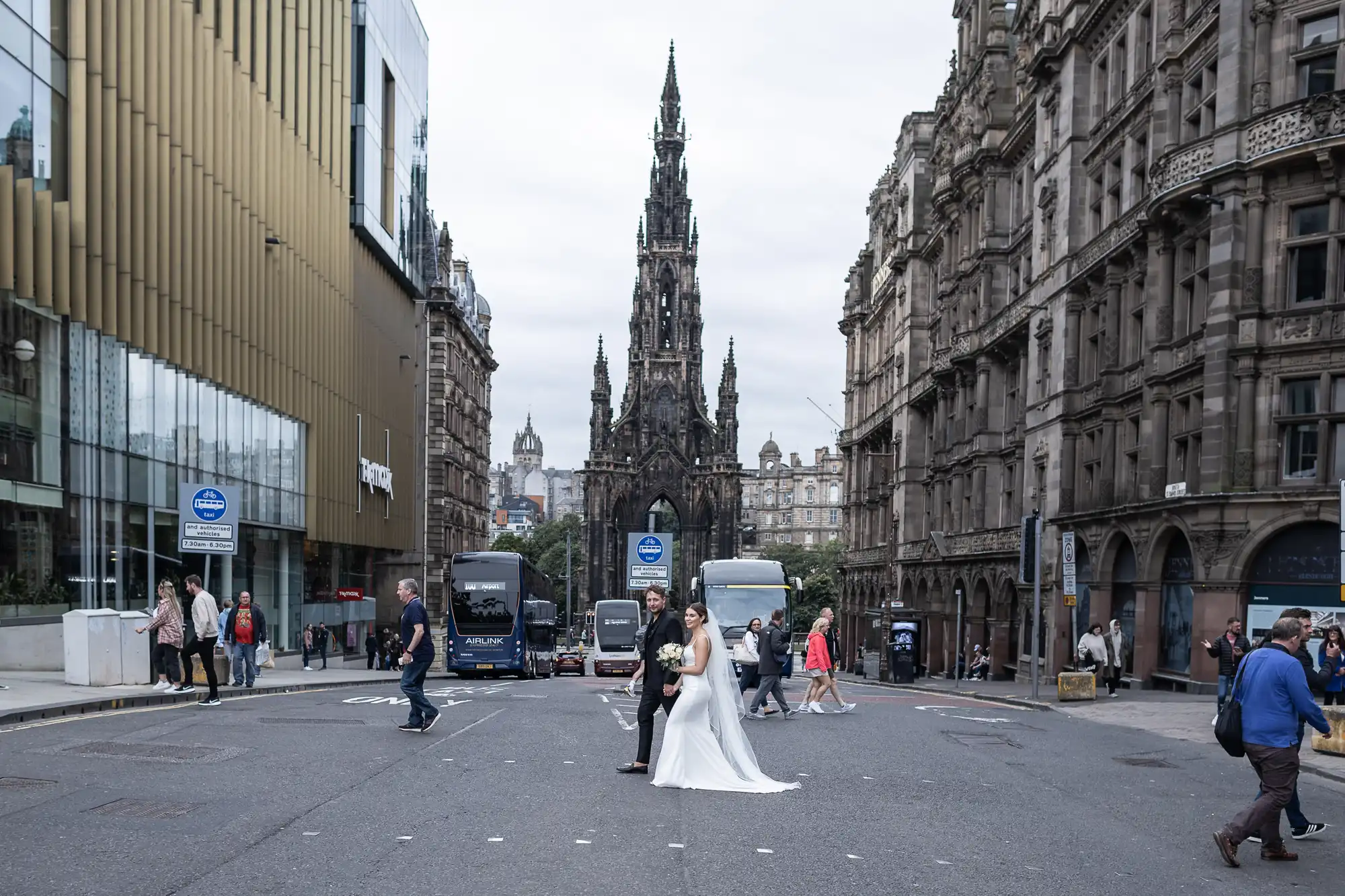 A bride and groom stand in the middle of a busy city street, posing for a photo with the scott monument in the background, surrounded by pedestrians and vehicles.