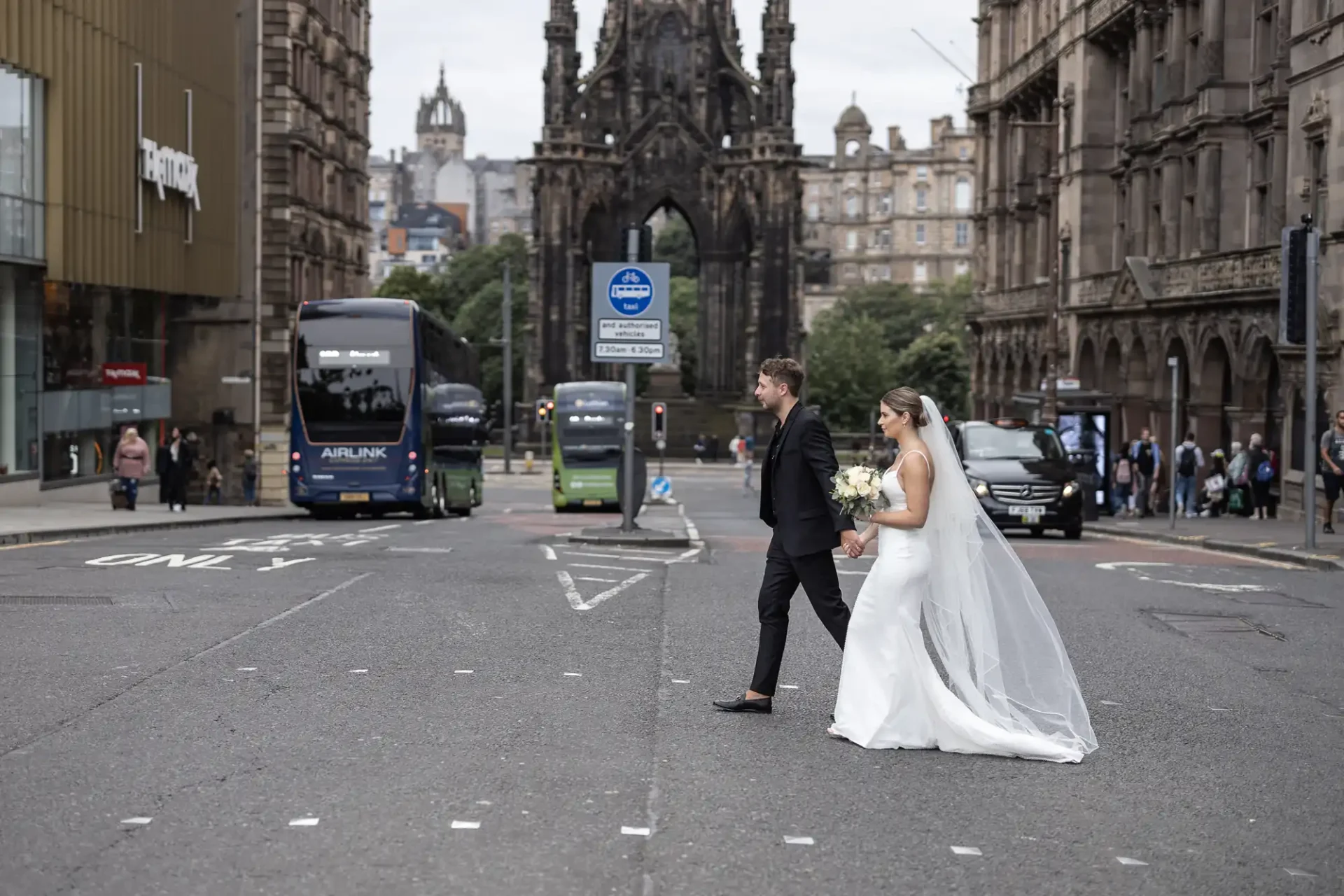 A bride and groom holding hands, crossing an urban street with a bus and classic architecture in the background.