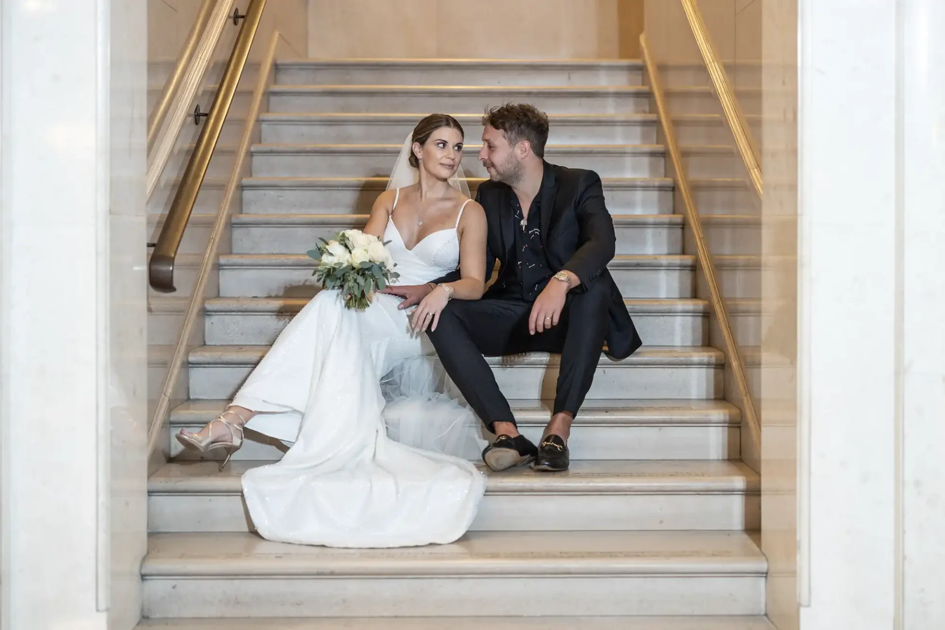 A bride in a white dress and a groom in a black suit sit closely on marble stairs, exchanging a loving gaze, with the bride holding a bouquet.