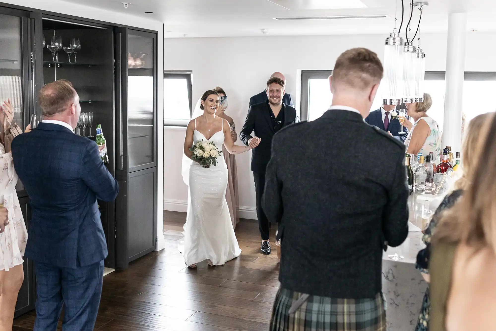 A bride and groom joyfully walk through a crowd of applauding guests in a modern, well-lit room.