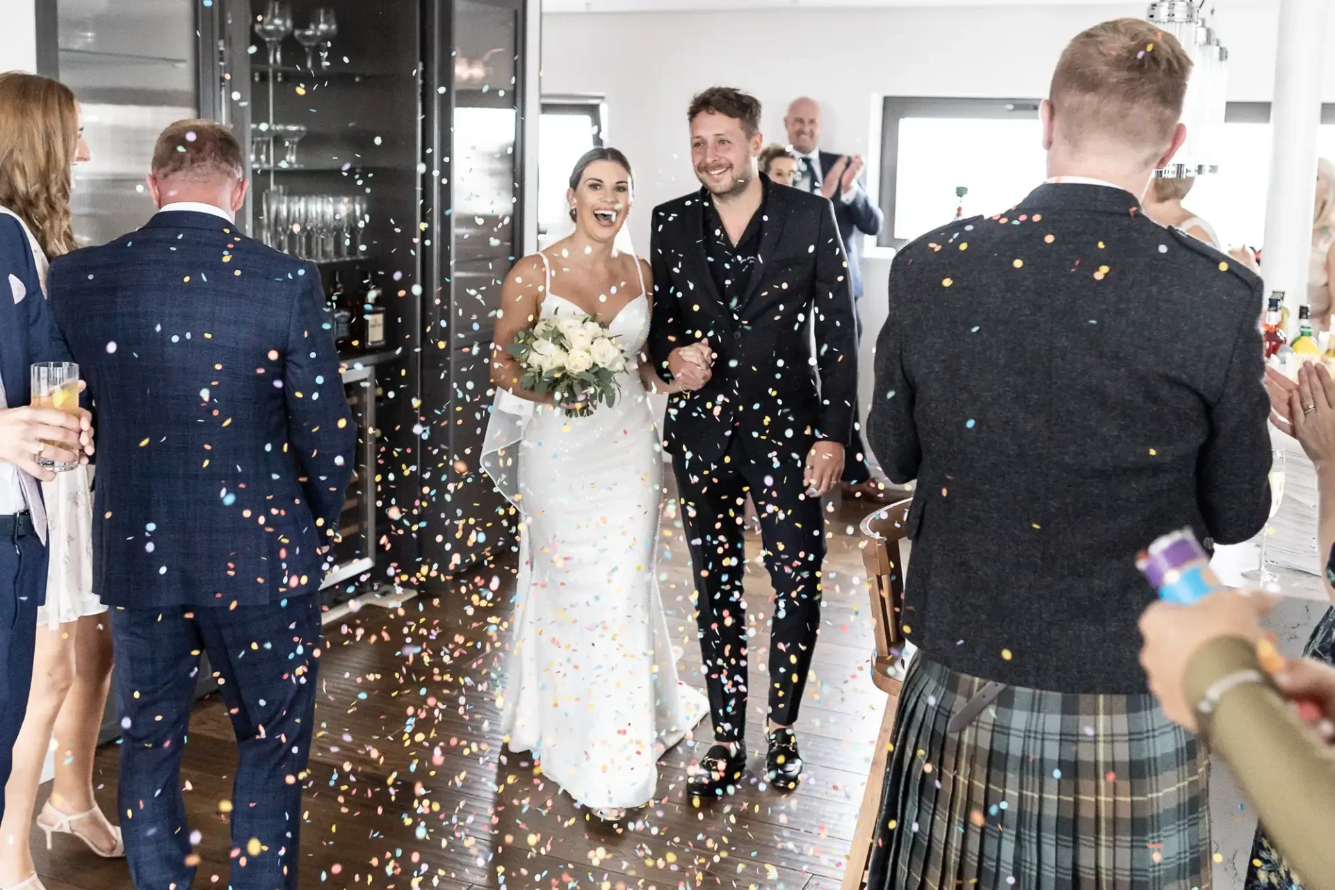 A newlywed couple joyfully walking through a shower of confetti, greeted by guests in a bright, elegant room.