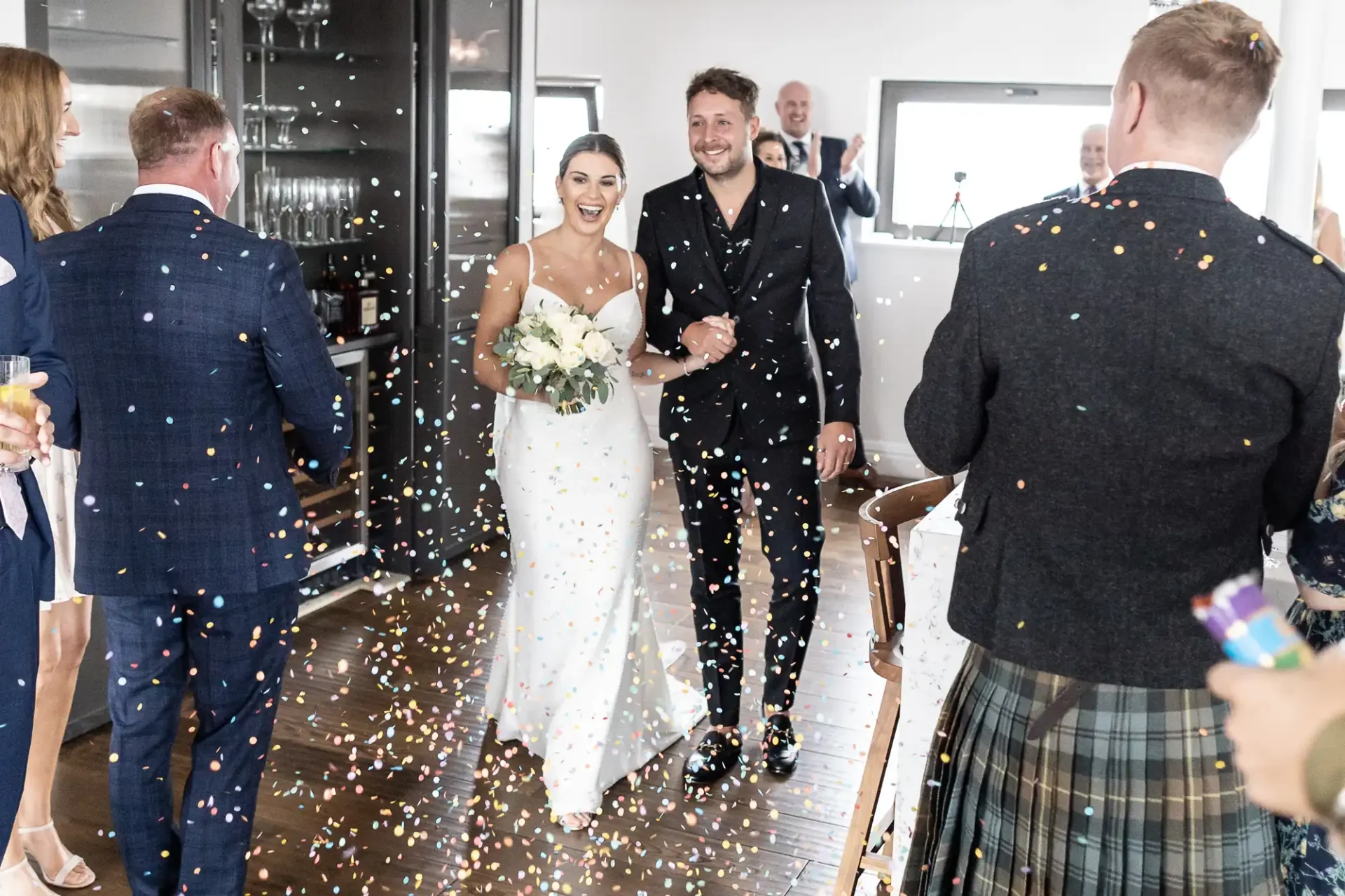 A newlywed couple walks happily through a crowd throwing confetti, smiling broadly in a stylishly decorated room.