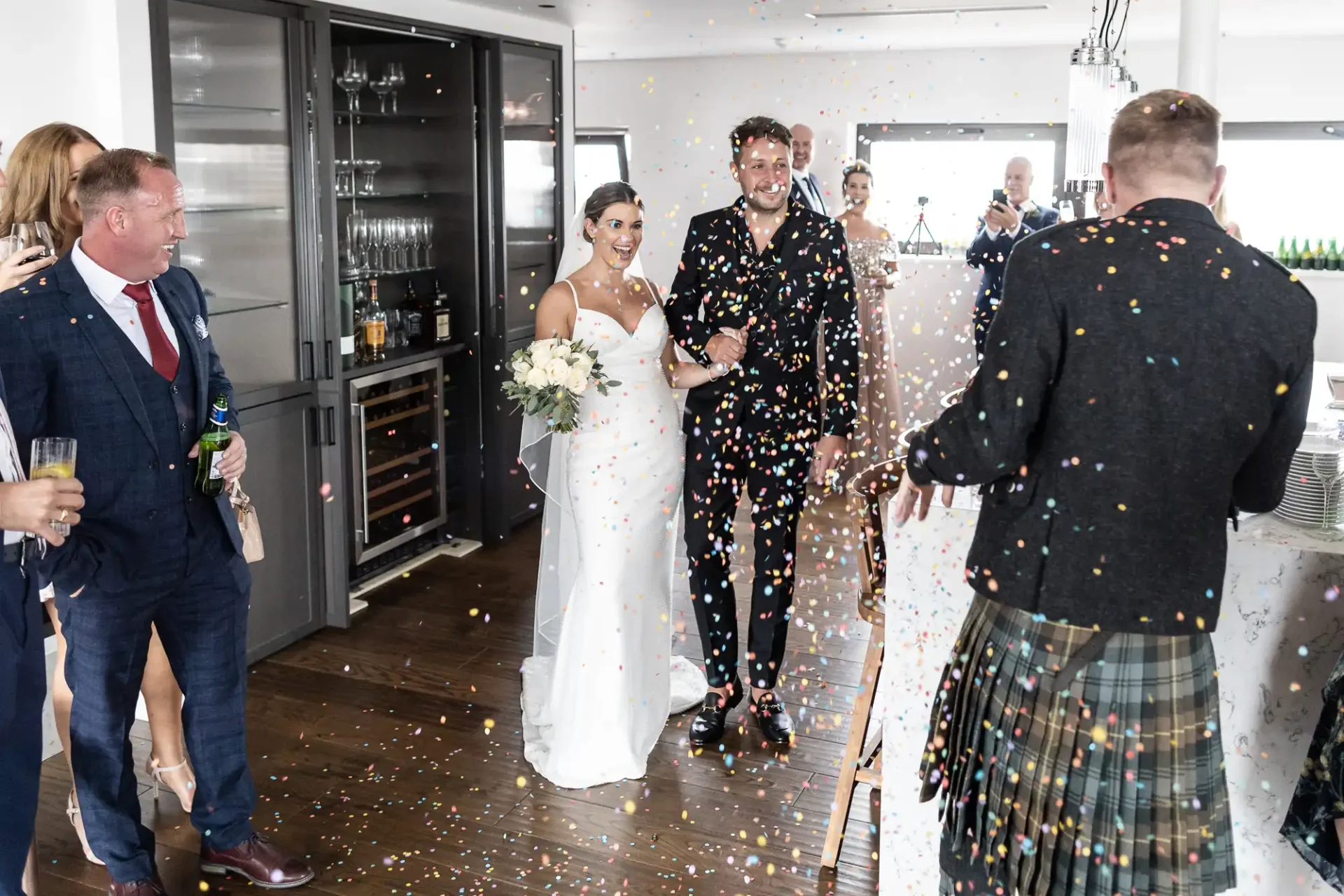 A newlywed couple walks through a scattered confetti shower, smiling, with guests observing in a brightly lit room.