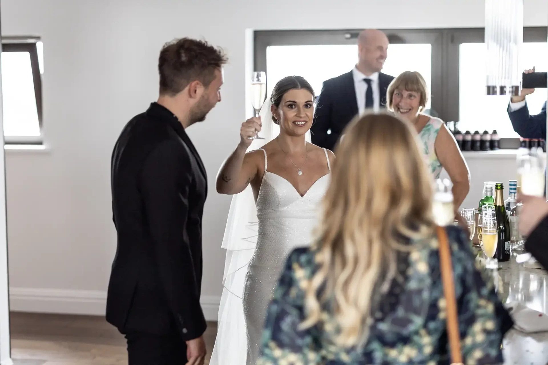 A bride holding a champagne glass greets a guest at a reception, with a smiling groom and other guests nearby in an elegant room.