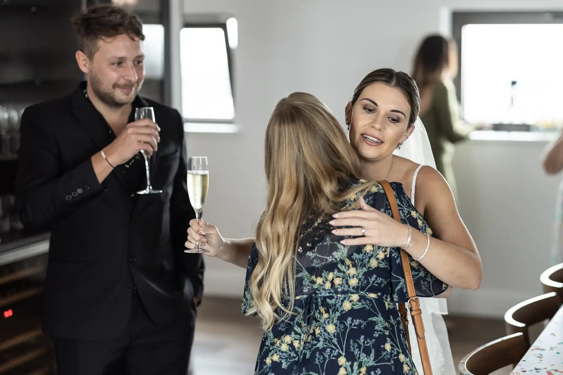 Two women and a man in semi-formal attire holding champagne glasses at a social gathering, engaging in a friendly hug and conversation.
