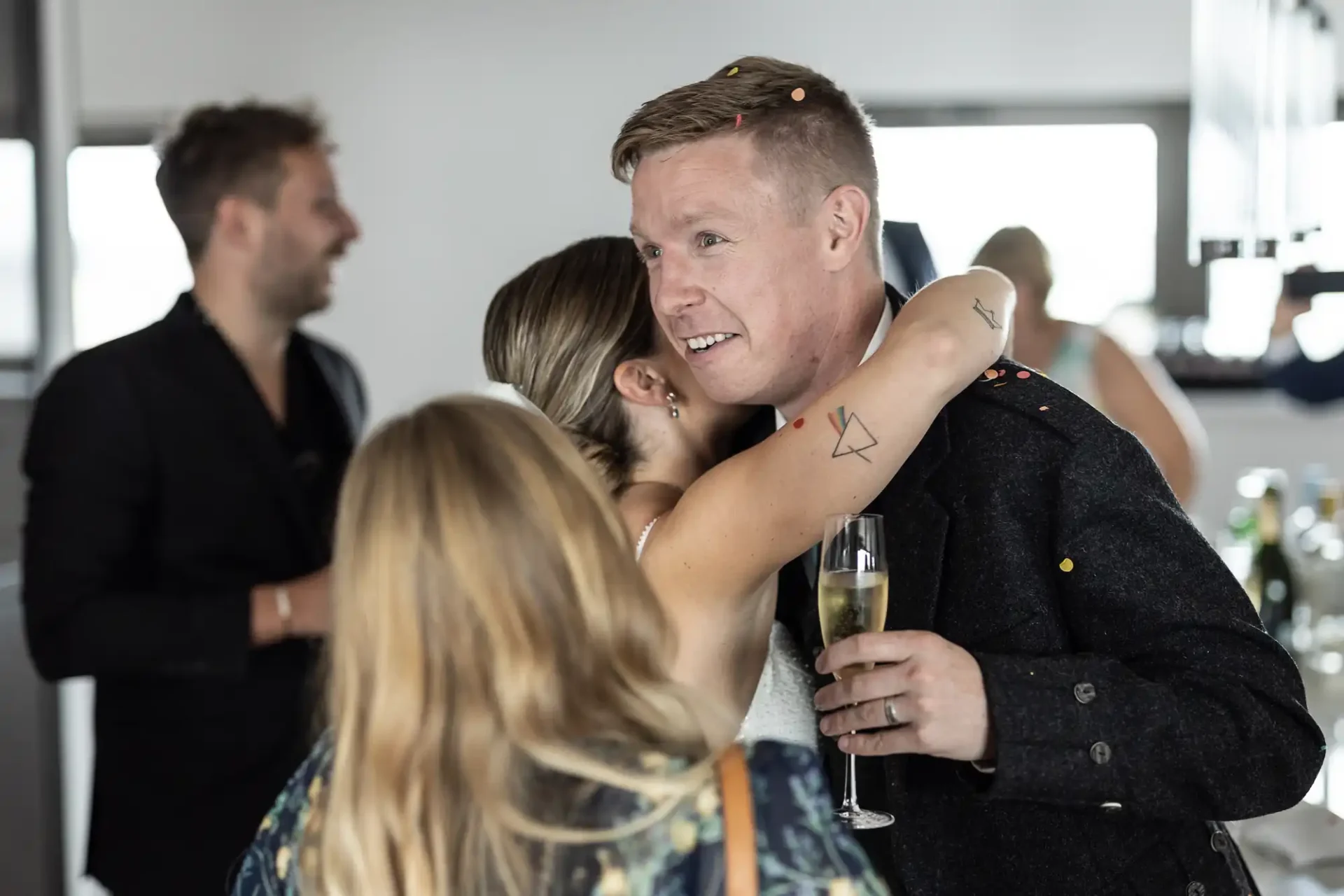 Man with a tattoo on his arm holding a champagne glass while greeting guests at a social gathering indoors.