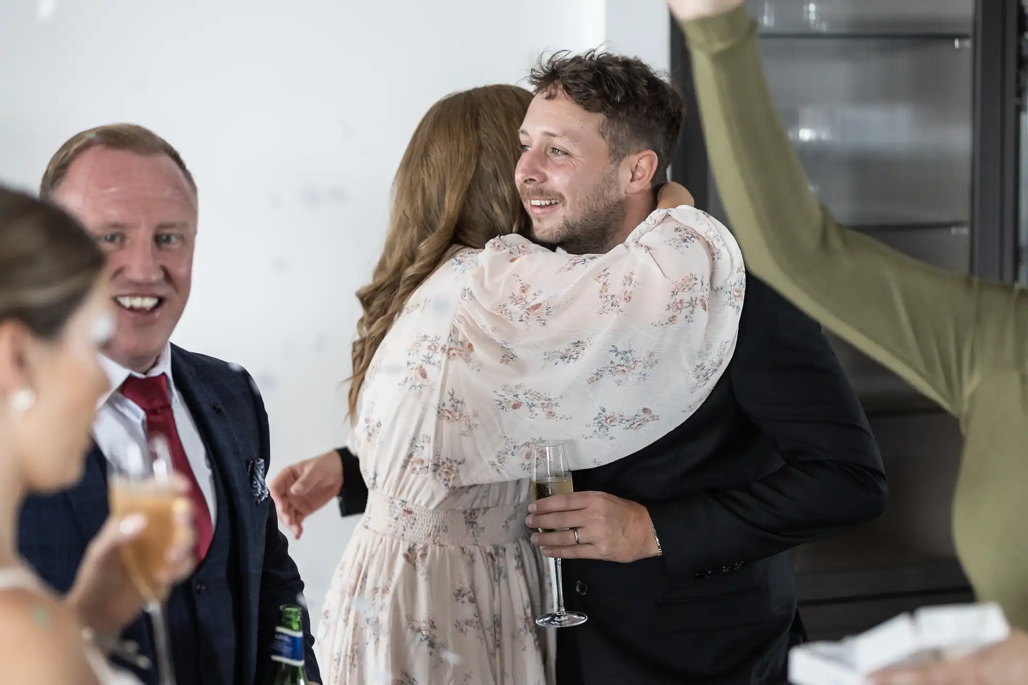 A man hugging a woman at a social gathering while holding a drink, with two other men smiling and interacting in the background.