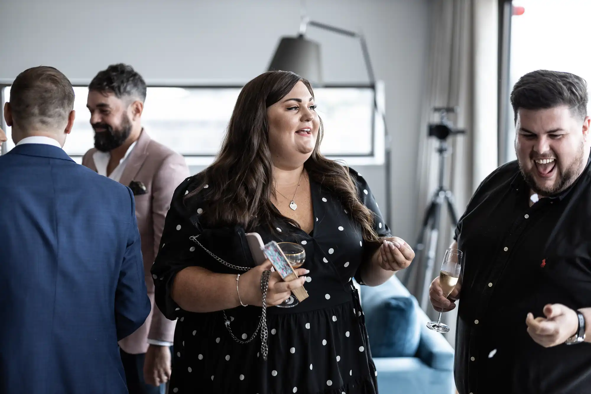 A group of adults socializing at an indoor event, with a woman in a polka dot dress holding a wine glass and smiling while talking to a man laughing beside her.