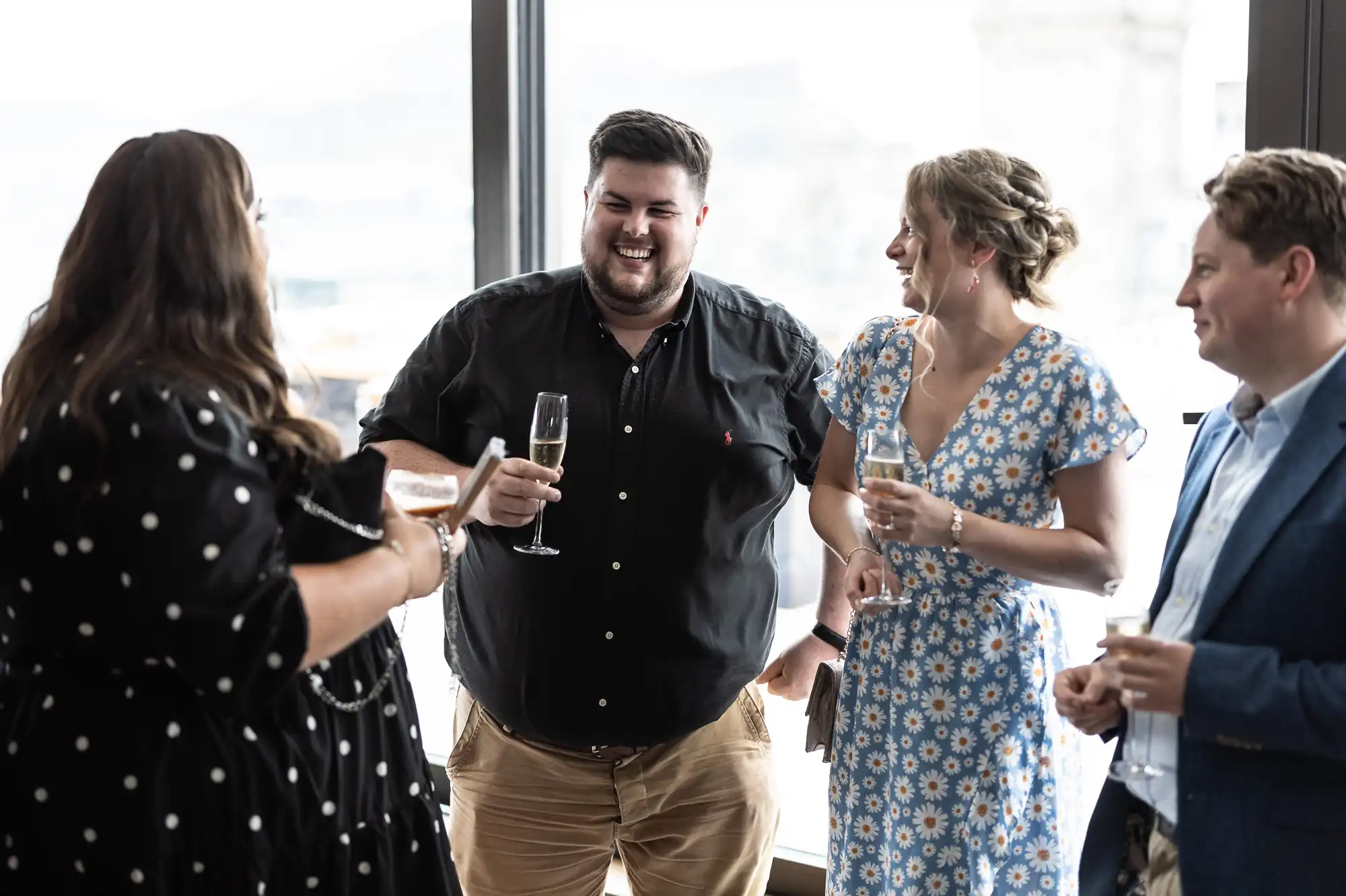 Group of four adults laughing and conversing at a social gathering, holding wine glasses, in a room with large windows.