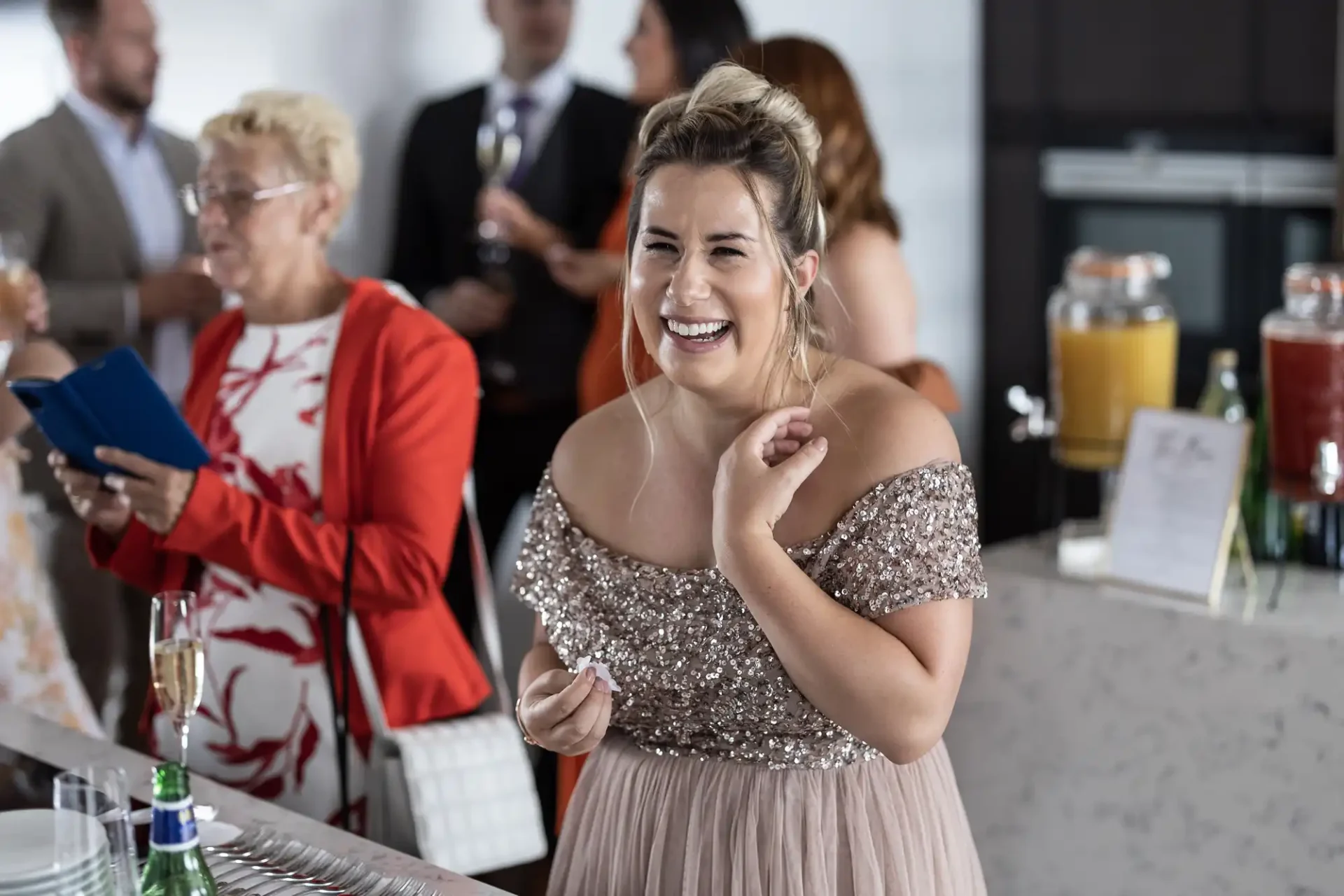 A woman in a glittery dress laughs joyfully at a social event, holding a clutch, with other guests and a drink station in the background.