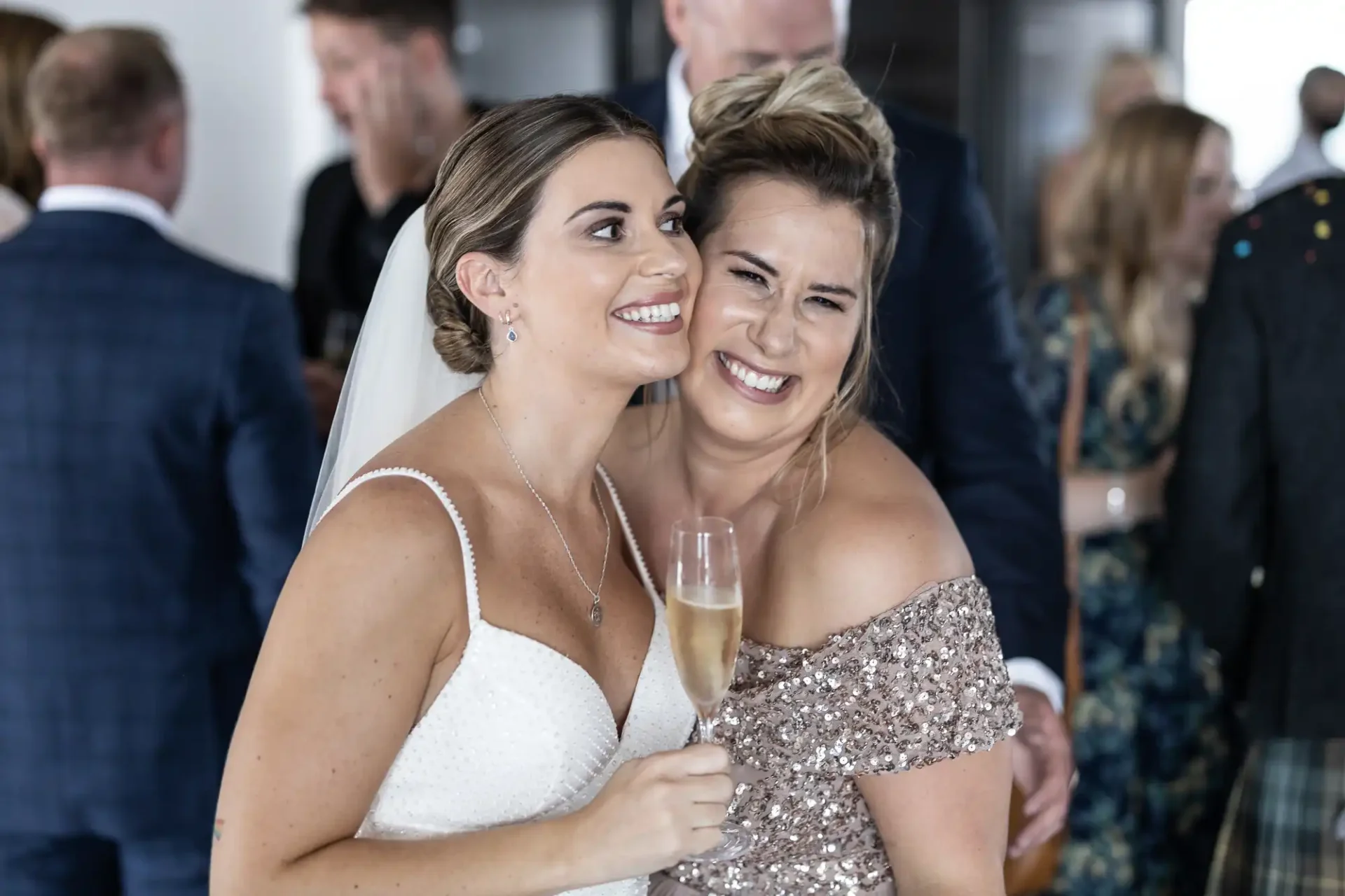 Two women smiling and embracing at a wedding celebration, one in a white bridal gown and the other in a sequined dress, holding a champagne flute.