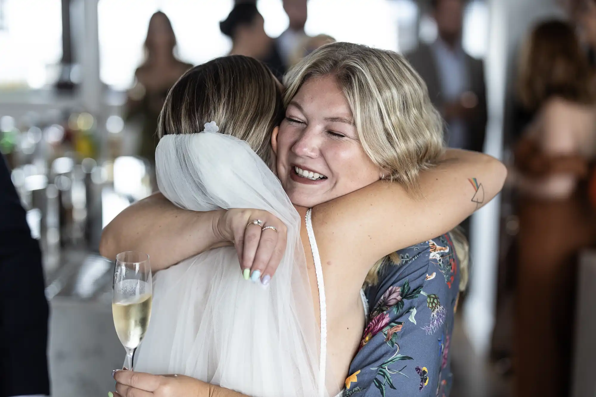 Two women embracing and smiling joyfully at a social gathering, one holding a glass of champagne.