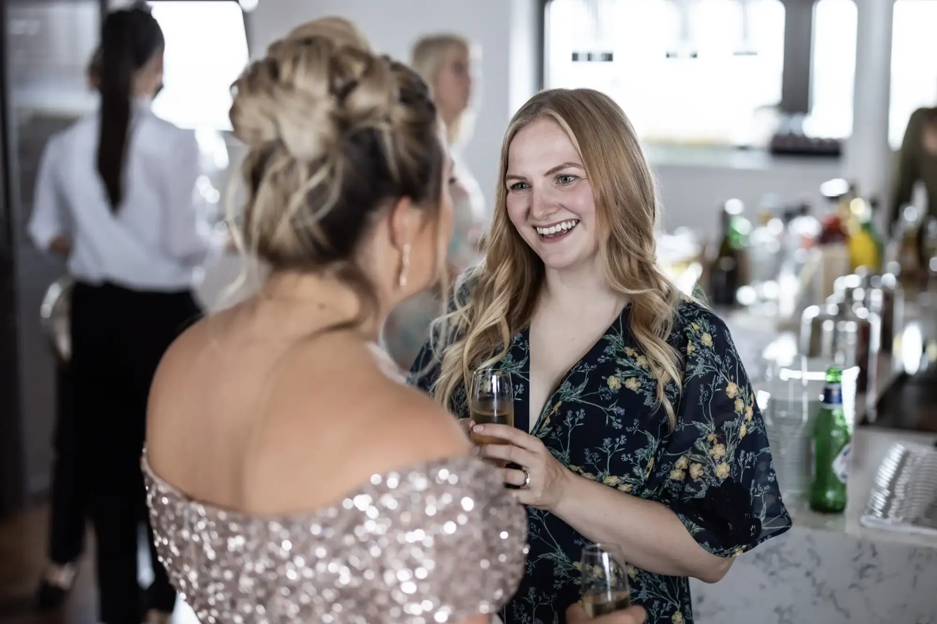 Two women, one in a floral dress and the other in a sequined top, conversing happily with drinks in hand at a social event.
