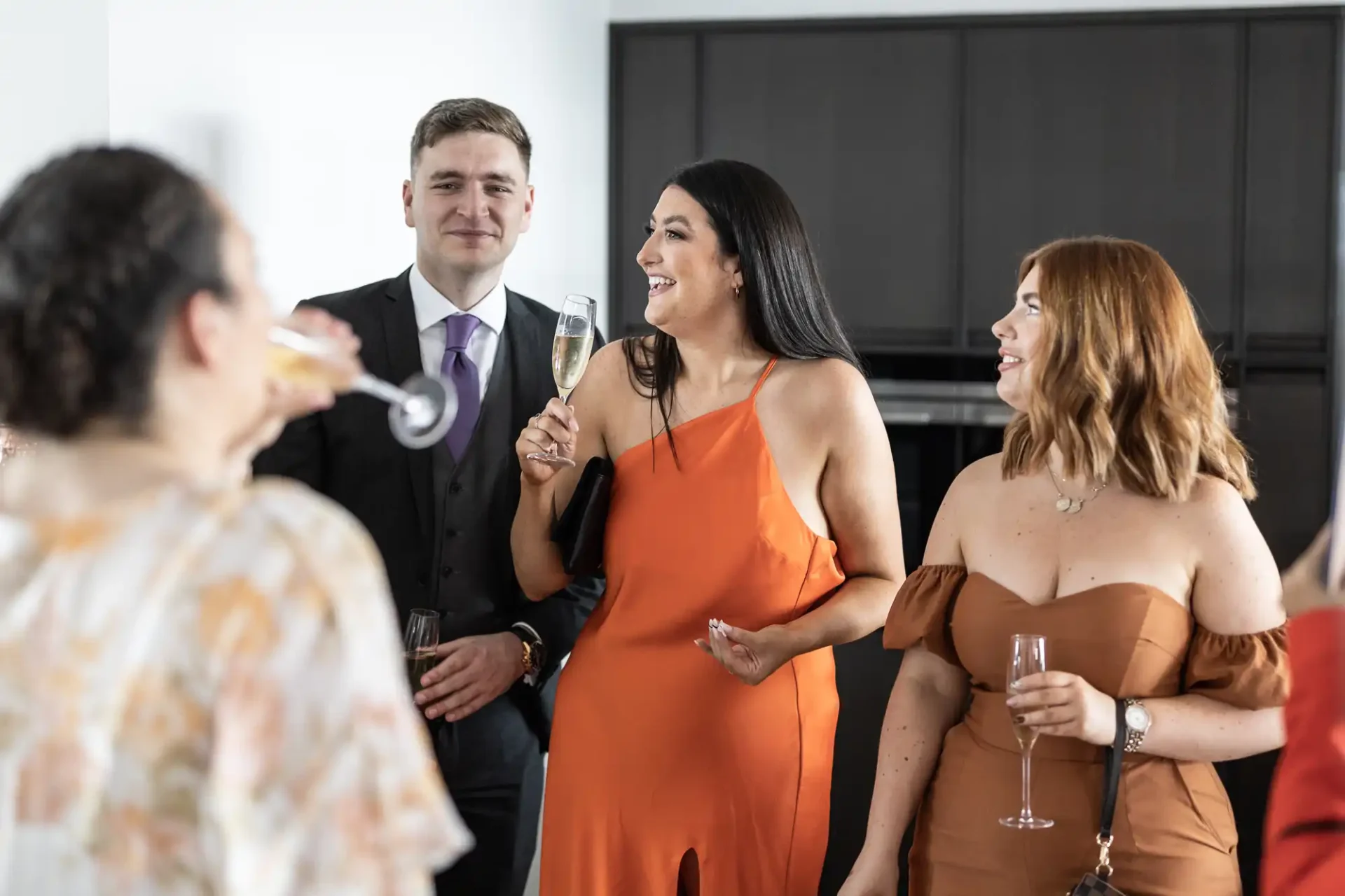 A group of people smiling and socializing at a party, holding champagne flutes, with a woman in an orange dress speaking animatedly.